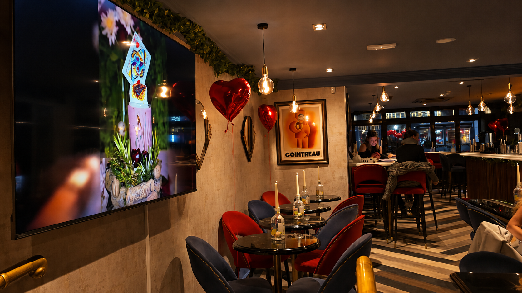 A cozy Bar with dim lighting, decorated with red heart-shaped balloons and hanging Edison bulbs. There are small tables with candles, velvet chairs, framed artwork on the wall, and a large screen displaying a photo of a plant with spring flowers. 97