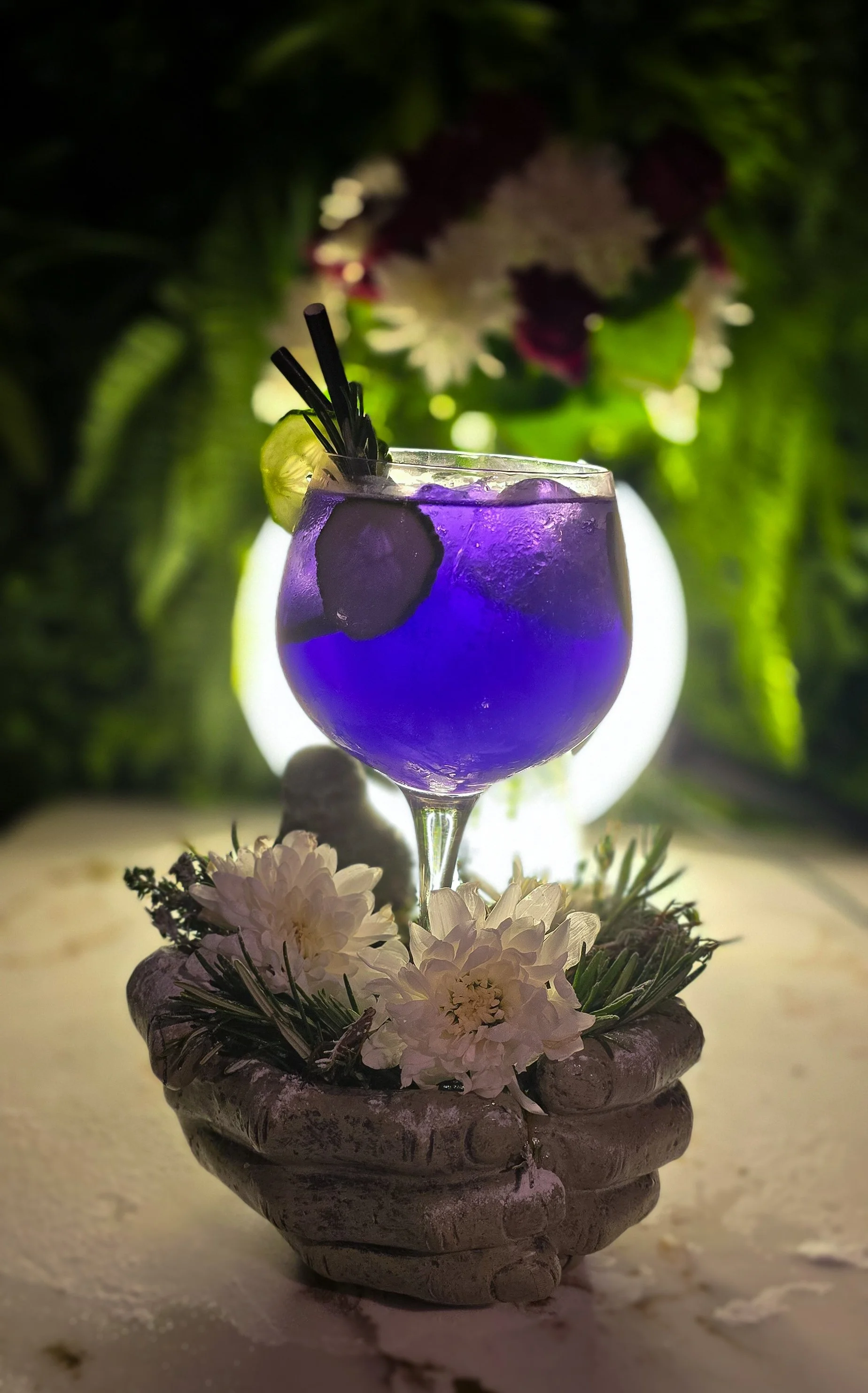 A vibrant purple cocktail with ice in a stemmed glass garnished with a lime wedge and black straws, placed in a decorative stone bowl filled with white flowers, set against a blurred green background with soft lighting. Signature cocktails at 97 bar.