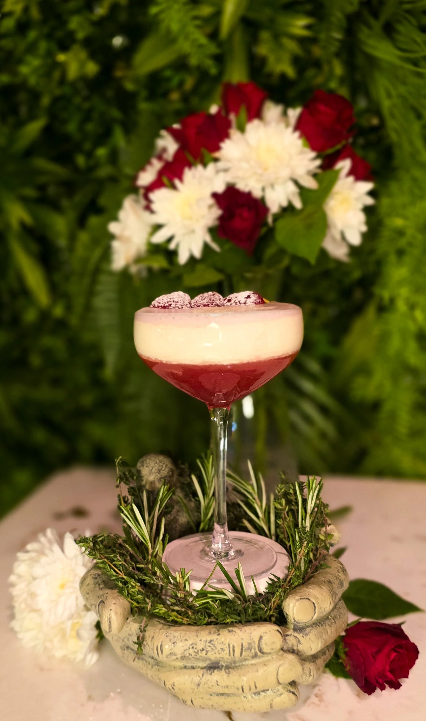 A pink cocktail garnished with raspberries and powdered sugar in a coupe glass, placed on a decorative stone centerpiece surrounded by greenery and flowers, with a backdrop of lush green foliage and white and red flowers. A signature cocktail at 97.