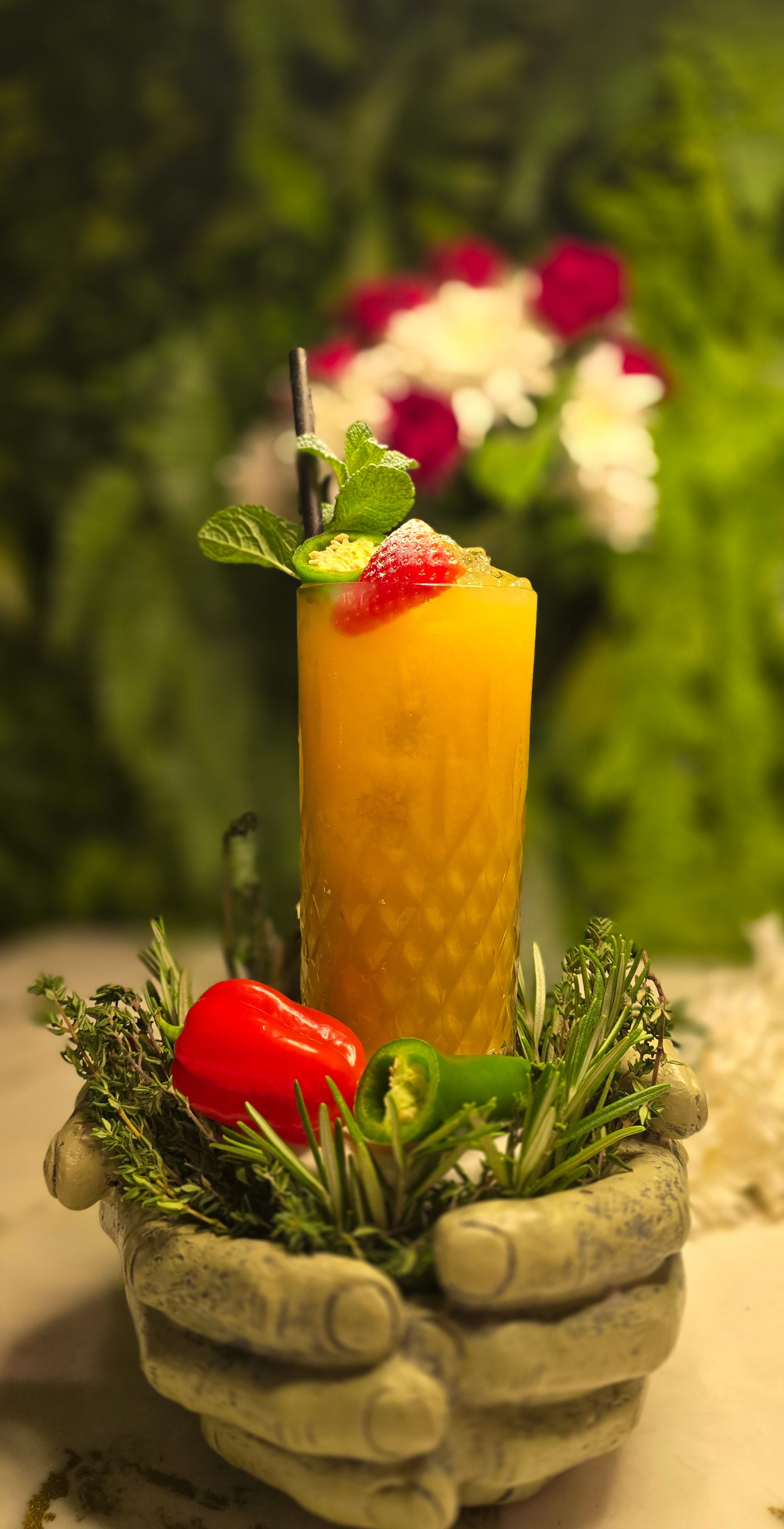 A tall glass of yellow-orange fruity cocktail garnished with a strawberry, mint leaves, and a slice of lime, placed in a container decorated with herbs, peppers, and flowers, with a blurred green background. Available at ninetyseven.