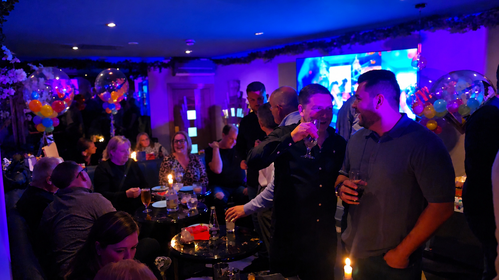 People socializing and celebrating at a party with colorful balloon decorations, dim lighting, and a large screen display in the background. Private hire event at ninety seven bar Sidcup Kent.