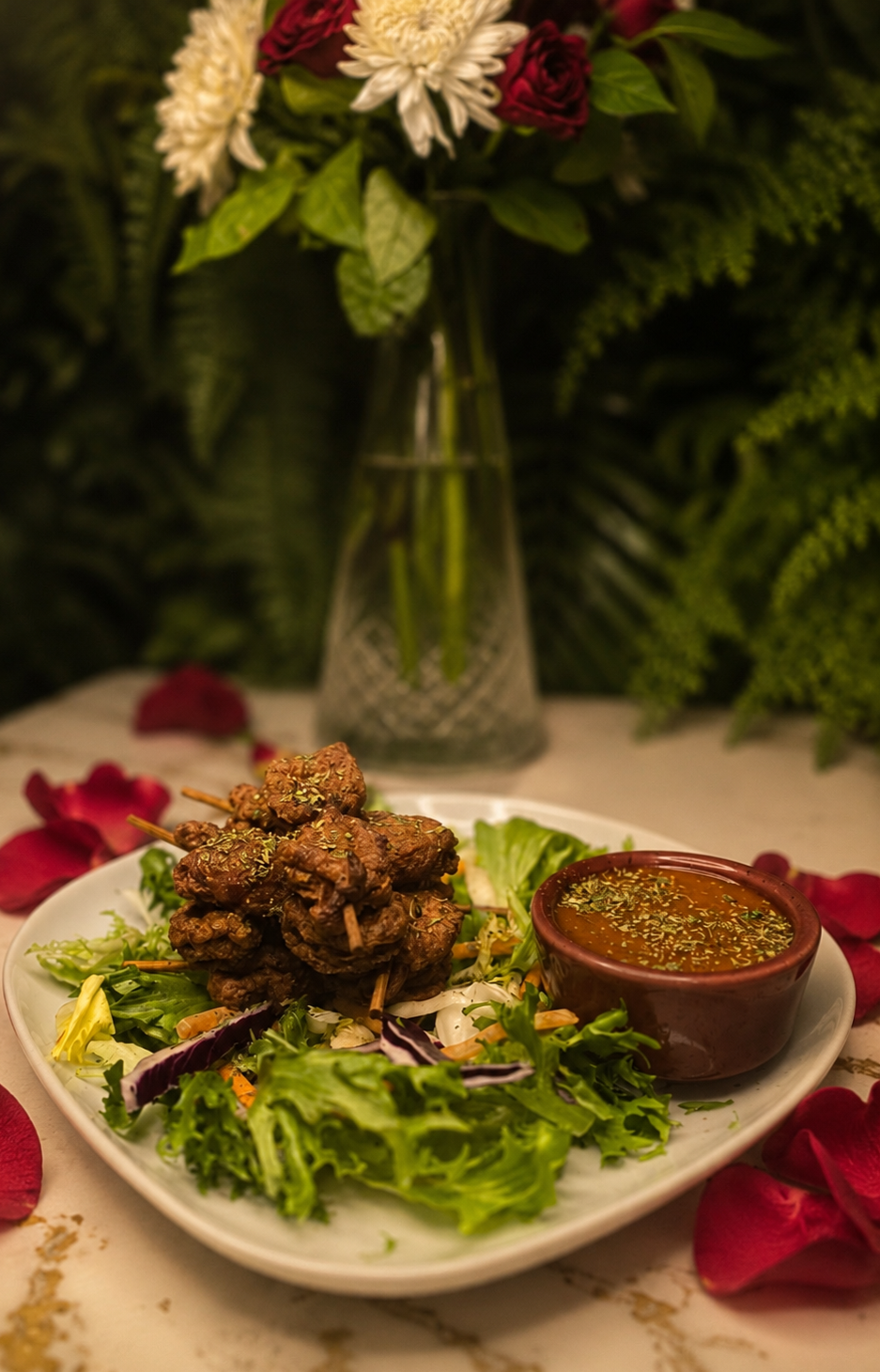 A plate with chicken skewers, a bowl of dipping sauce, and fresh salad greens. There are red rose petals scattered around the plate, and a vase with flowers in the background. Best seller at 97 Bar.
