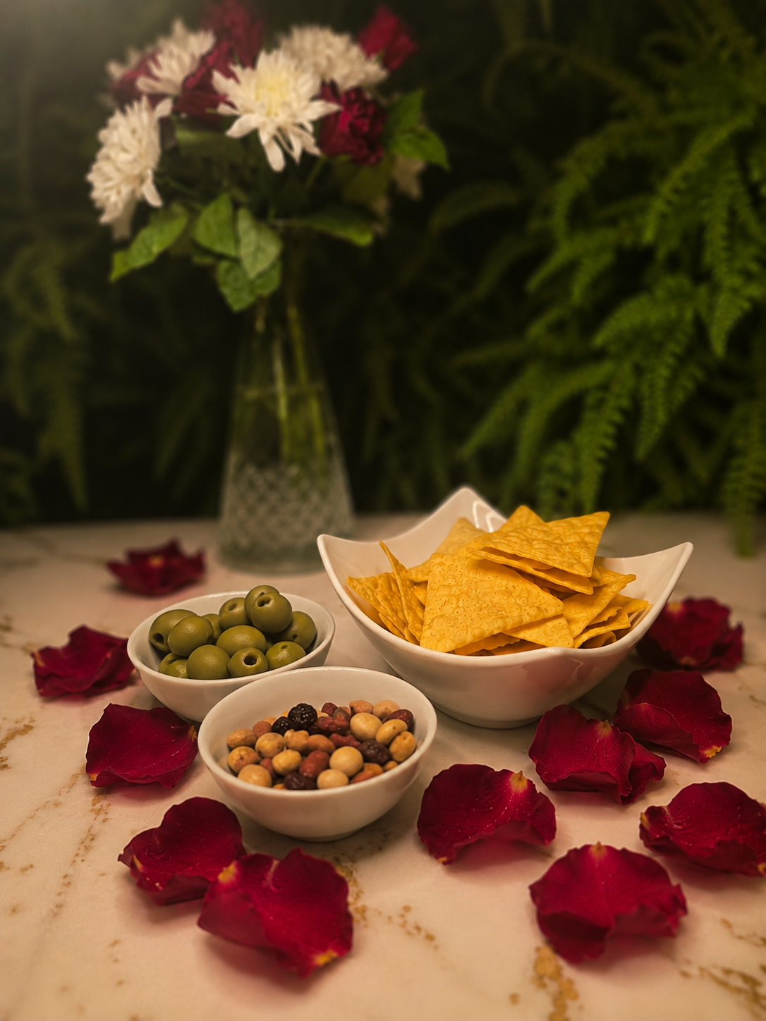 A table with bowls of green olives, mixed nuts, and cheese crisps, surrounded by red rose petals. In the background, a vase with white and red flowers in front of green ferns. Nibbles available at Ninetyseven bar Sidcup.
