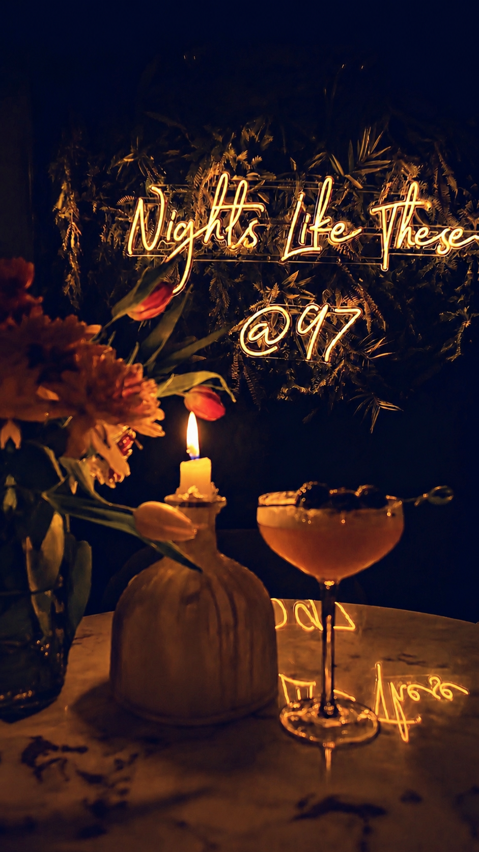 A romantic candle lit table with a vase of flowers, a lit candle, and a cocktail glass, with a neon sign in the background reading "Nights Like These @ 97". Ninety Seven bar, an atmospheric bar in Sidcup Kent.