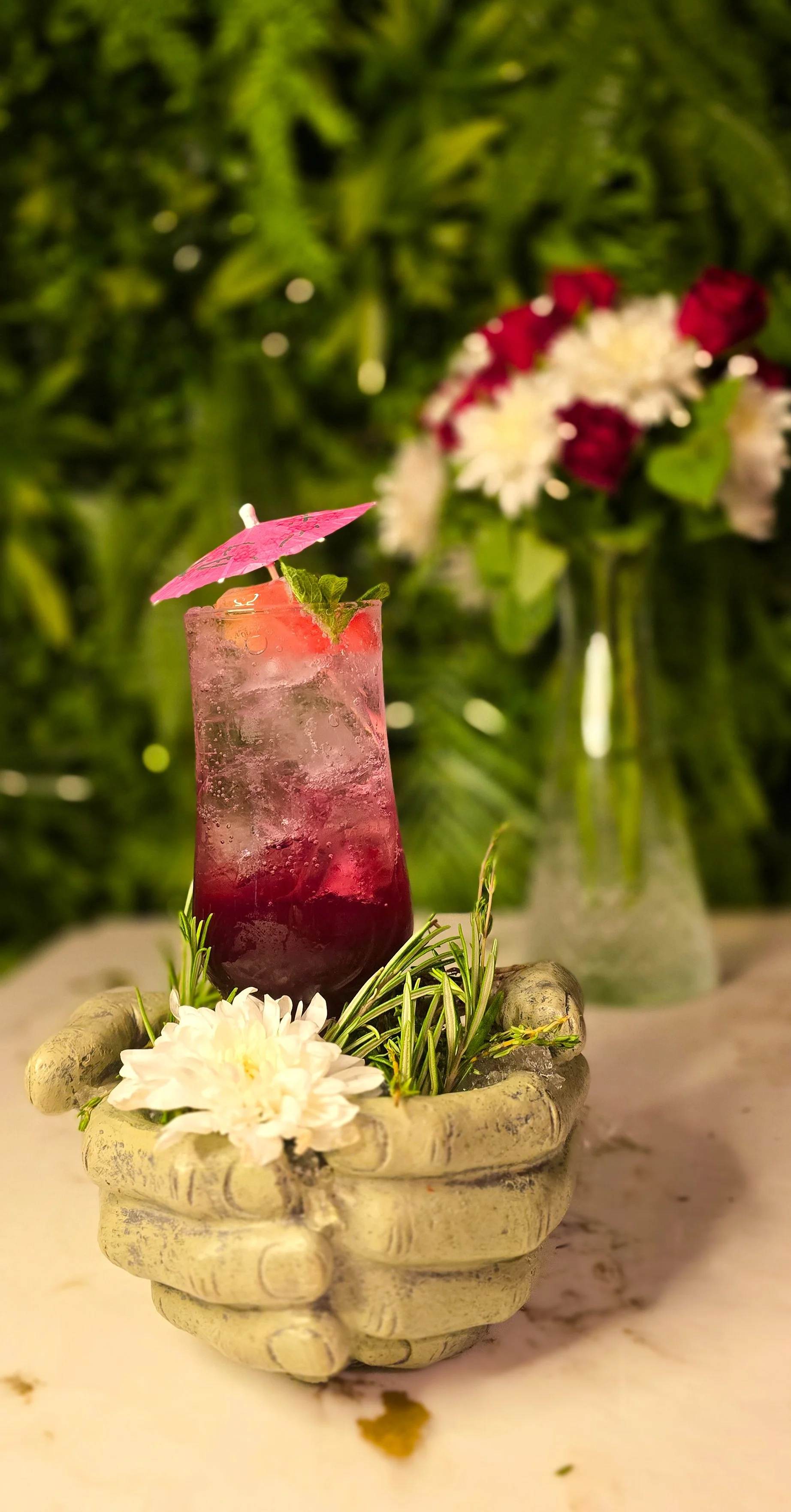 A tall glass with a pink and purple layered cocktail decorated with a pink paper umbrella, mint leaves, and a strawberry, with white flowers and greenery, against a background of blurred green plants and a vase with flowers. Ninety Seven Bar