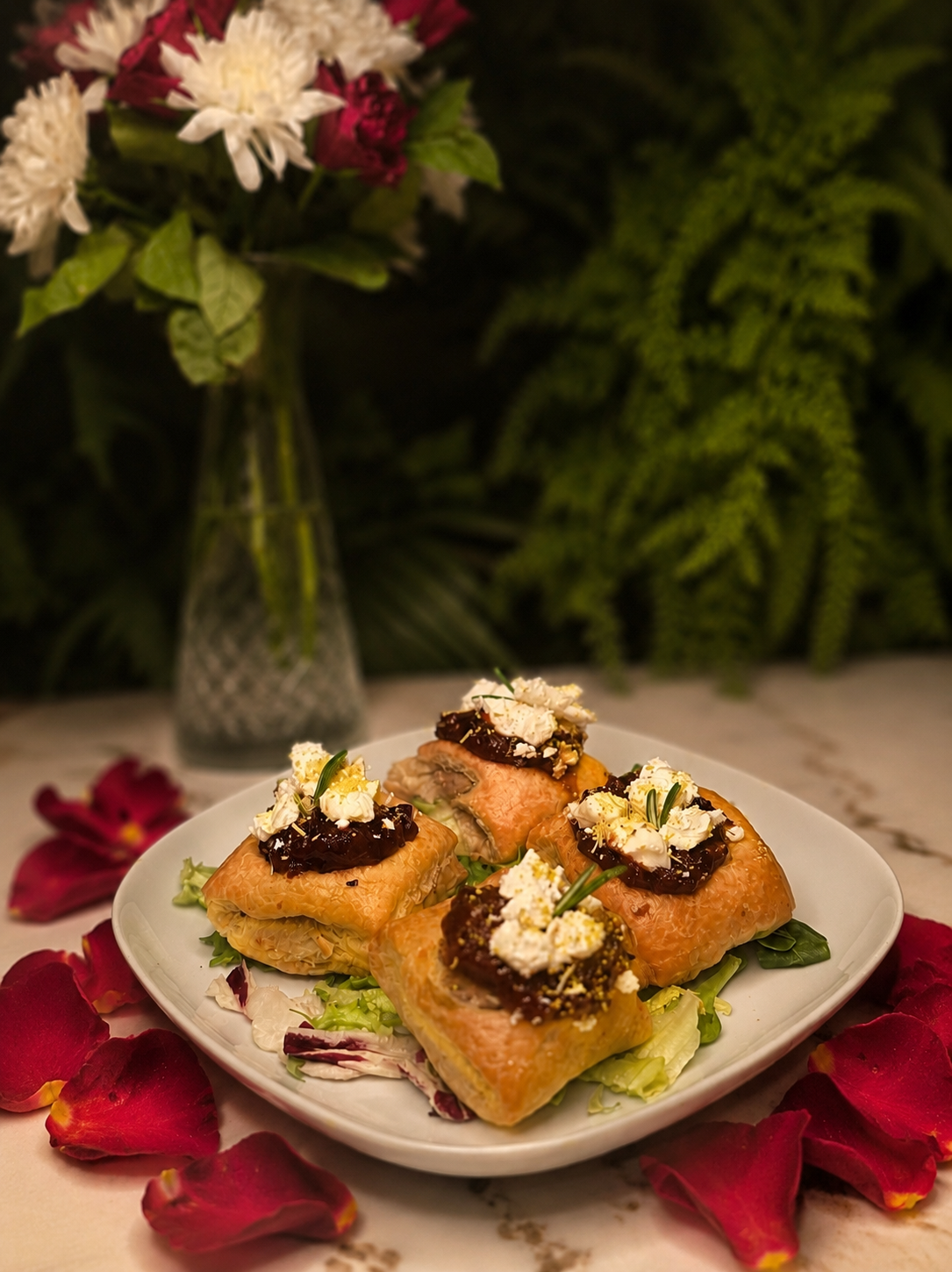 A plate of four puff pastry appetizers topped with cheese, jam, and herbs, garnished with white crumbled cheese and green herbs, all arranged on a bed of salad leaves, with rose petals scattered around the plate, and a vase with white and dark red flowers in the background.