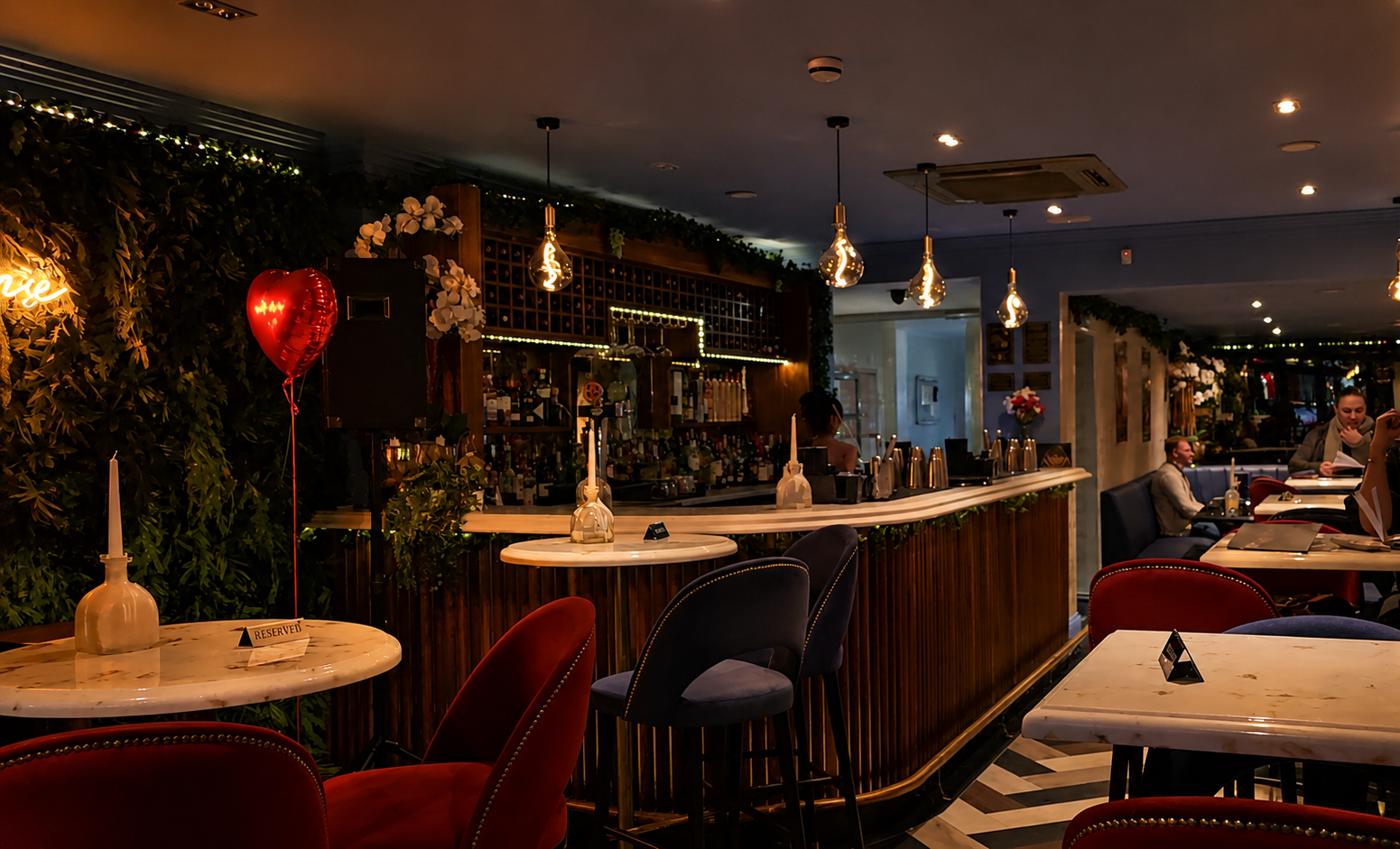 Interior of a cozy bar & lounge , Ninety Seven Sidcup, decorated with greenery, flowers, and warm lighting. Marble tables with reserved signs, velvet chairs, and a bar with various bottles and glasses in the background.