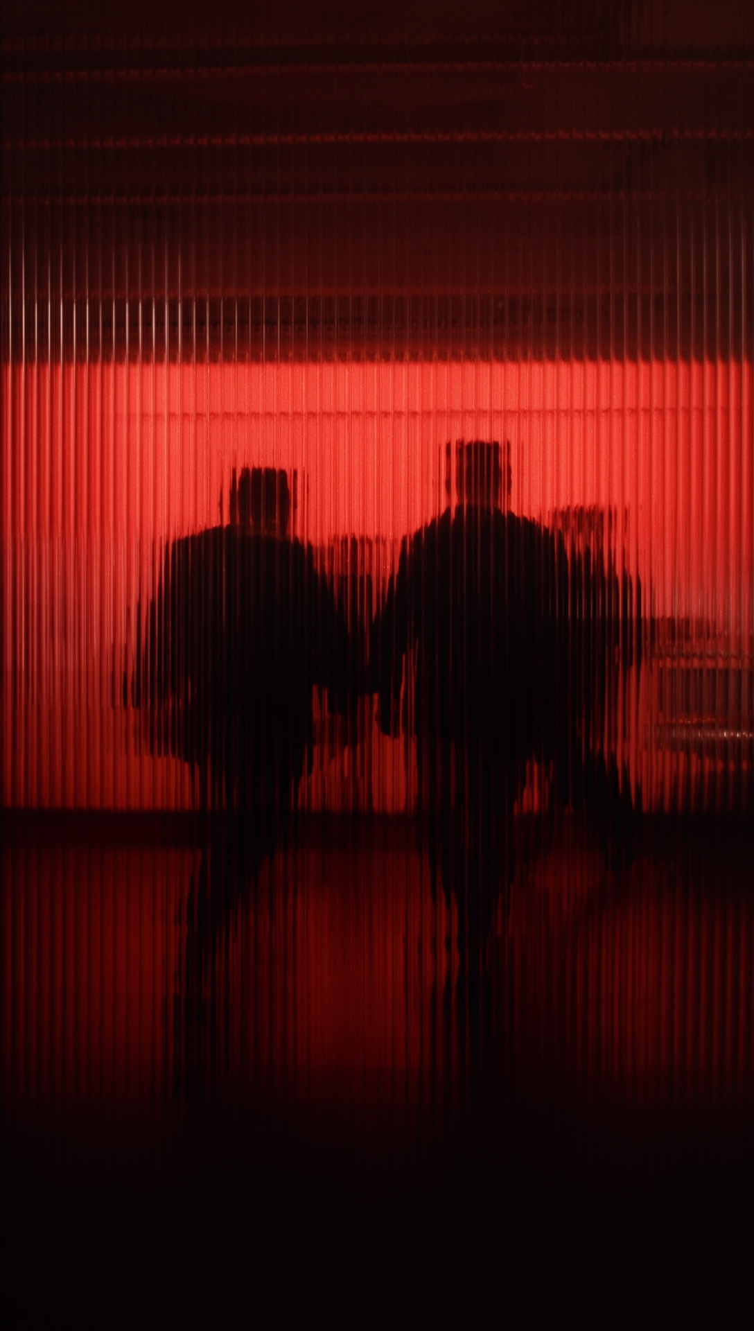 Siluetas de tres personas caminando juntas en un entorno oscuro con iluminación roja, vistas a través de una superficie con líneas verticales.