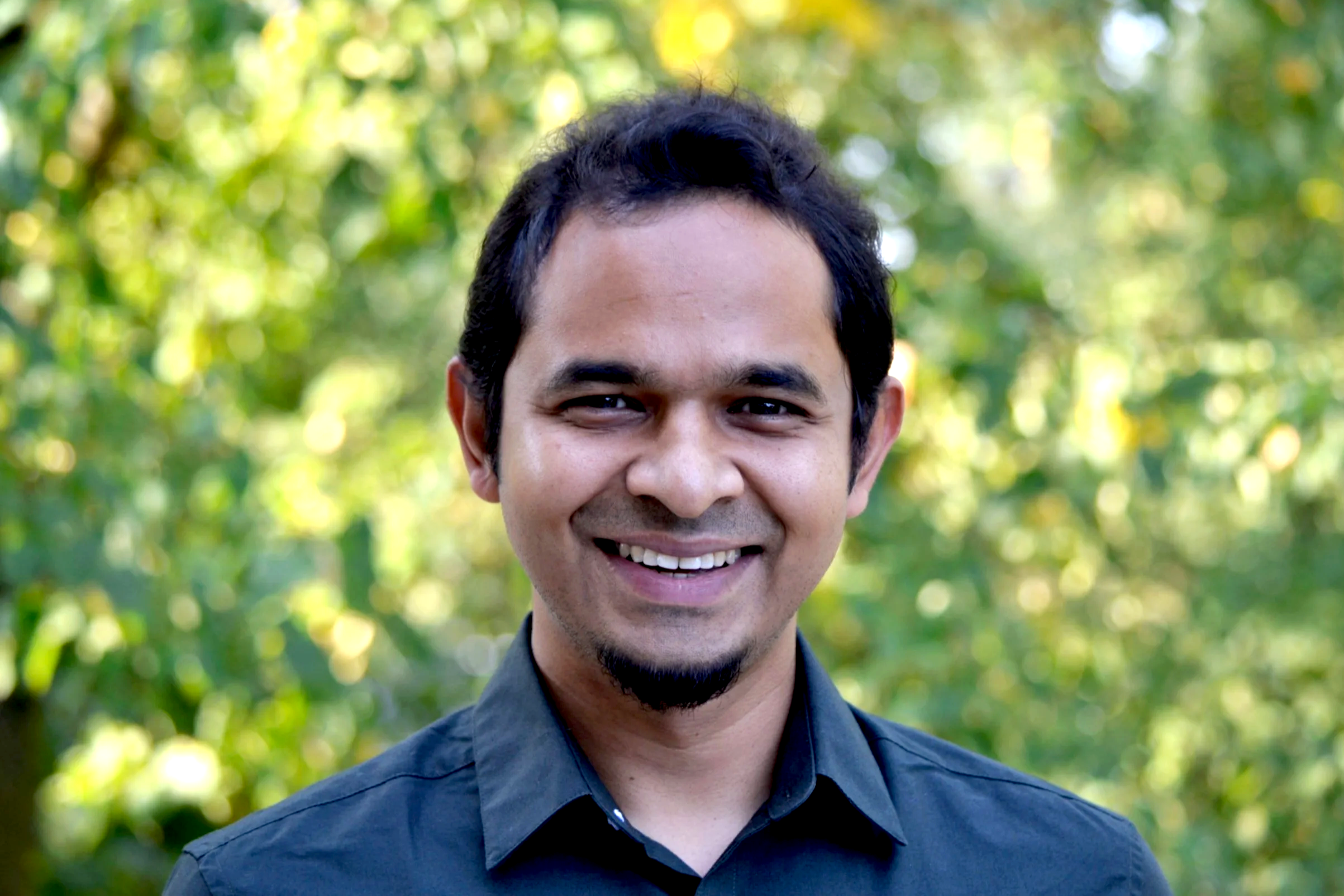 A smiling man with short dark hair, facial hair, and wearing a dark collared shirt standing outdoors with a blurred background of green and yellow foliage.