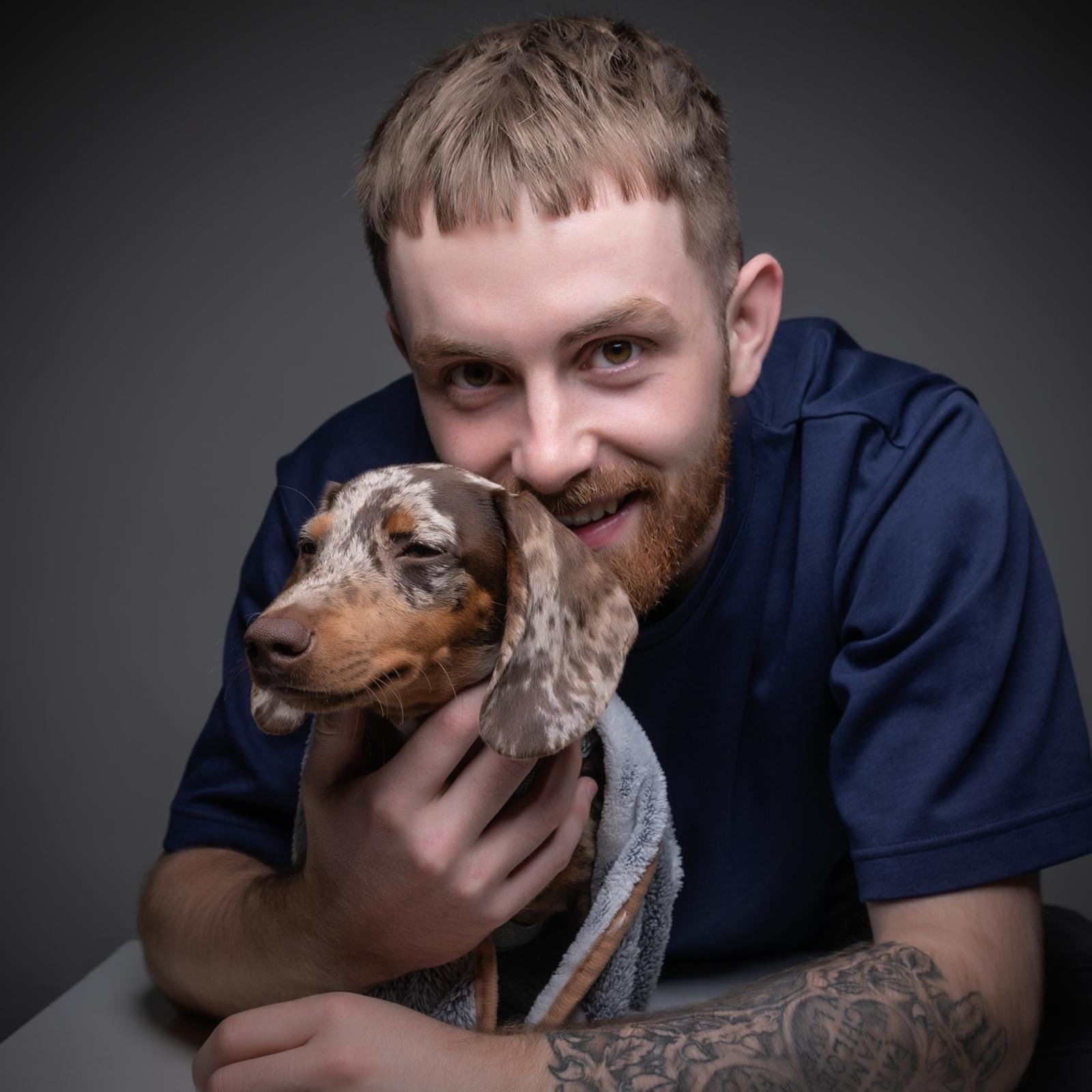 A young man with short brown hair and a beard is holding a Dachshund puppy with a speckled coat. The man is smiling slightly and wearing a dark blue shirt, and the background is dark gray.