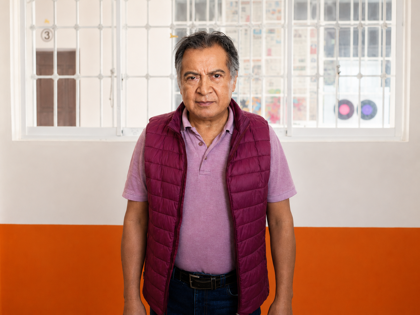 Hombre de edad media con cabello gris, vistiendo una camiseta lavanda y una chaqueta de rompevientos vino, de pie en una habitación con ventanas y rejas blancas, paredes blancas y una franja naranja en la pared.