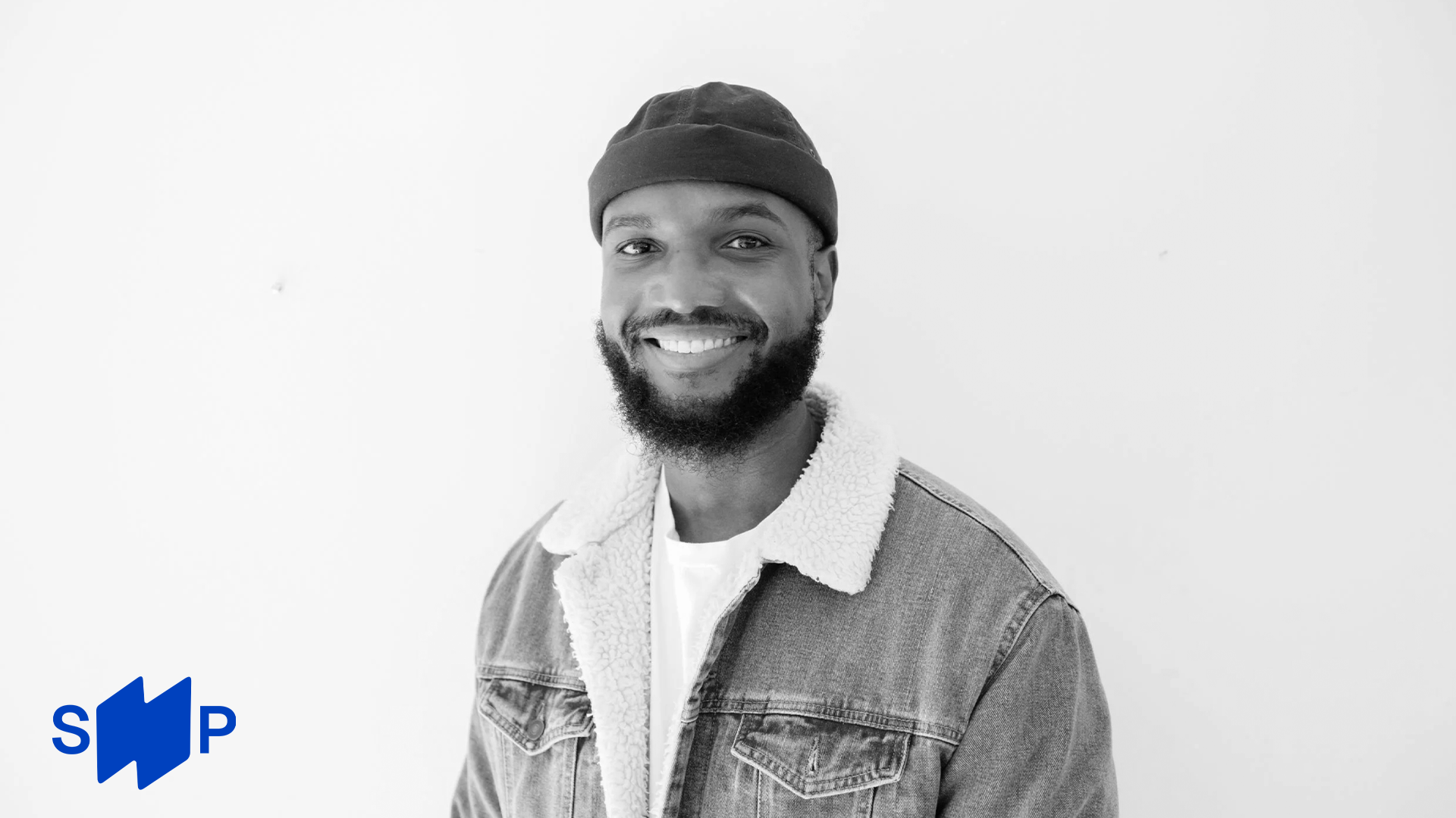 Black and white portrait of a smiling African American man wearing a beanie and denim jacket with sherpa lining, standing against a plain white background.