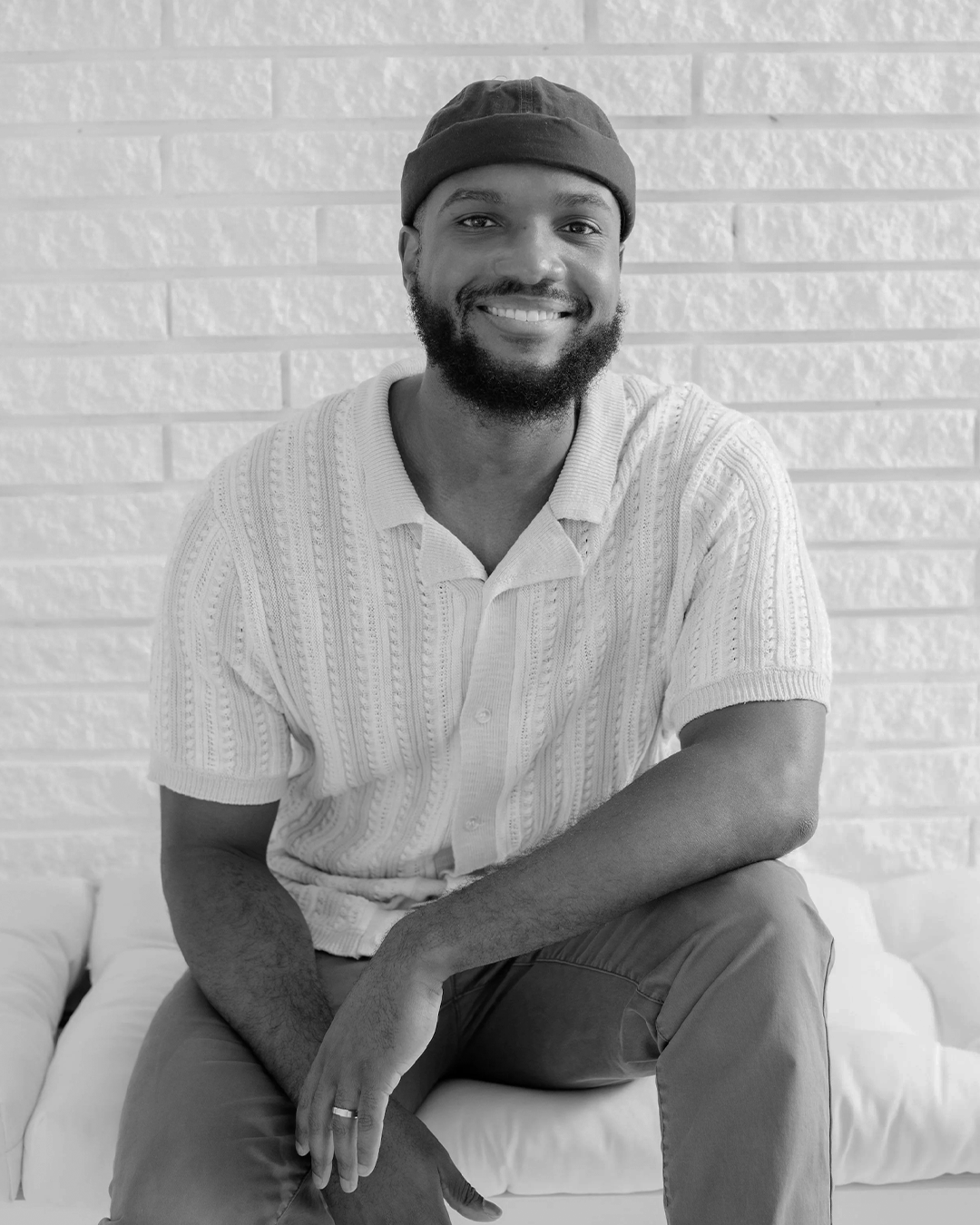 A smiling man with a beard wearing a hat and a short-sleeved button-up shirt sitting against a brick wall.