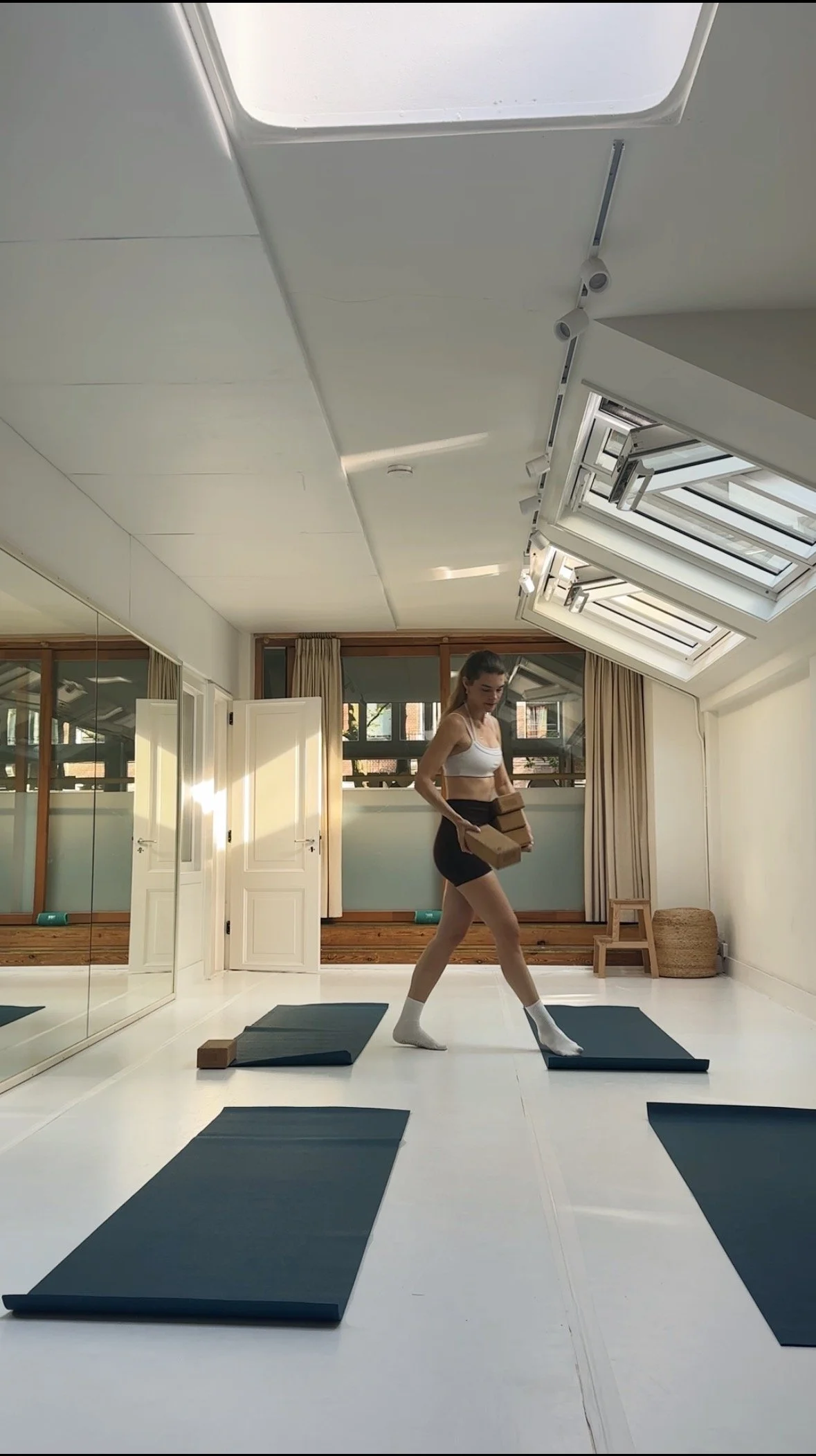 A woman in workout clothes standing on a yoga mat in a bright room with large windows, holding yoga blocks, and surrounded by other yoga mats.