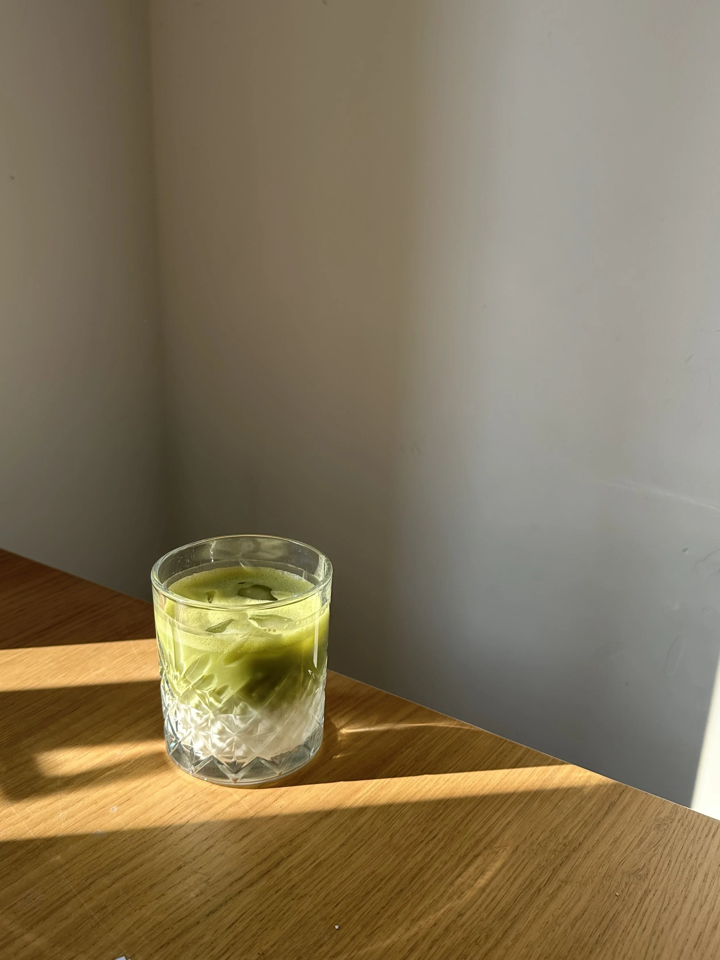 A glass with a green drink on a wooden table, with a plain light-colored wall in the background.
