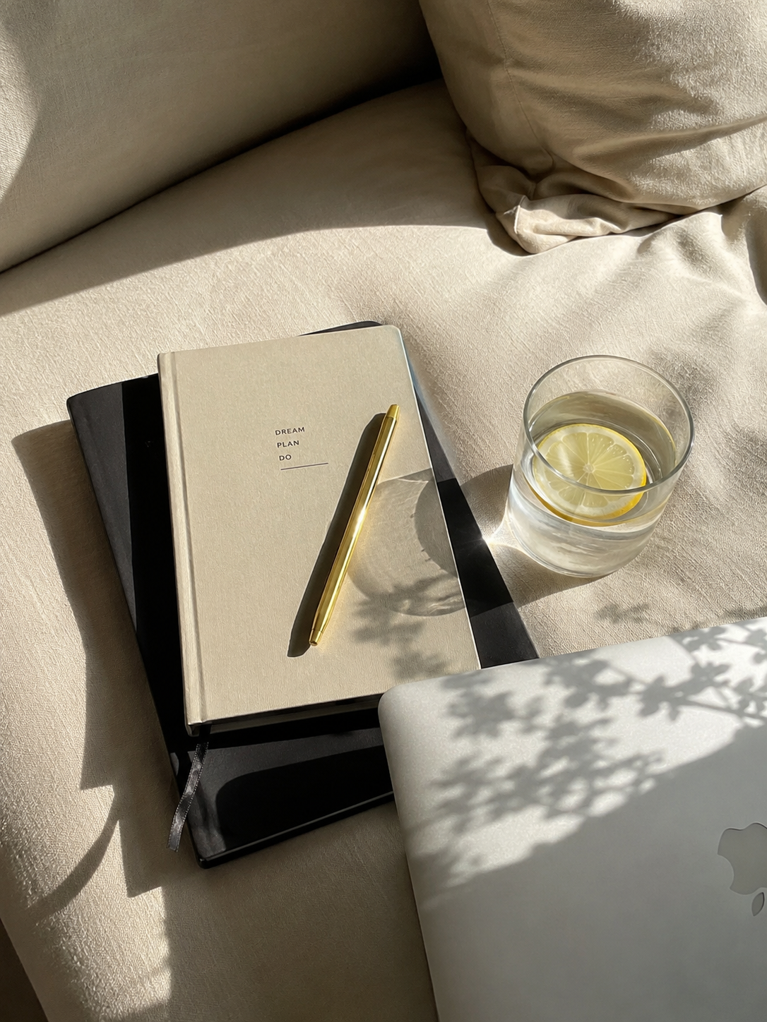A beige couch with a beige pillow, a stack of three notebooks (black, white, and darker), a gold pen on the white notebook titled "DREAM PLAN DO," a glass of water with lemon slices, and a closed silver MacBook laptop with an apple logo.