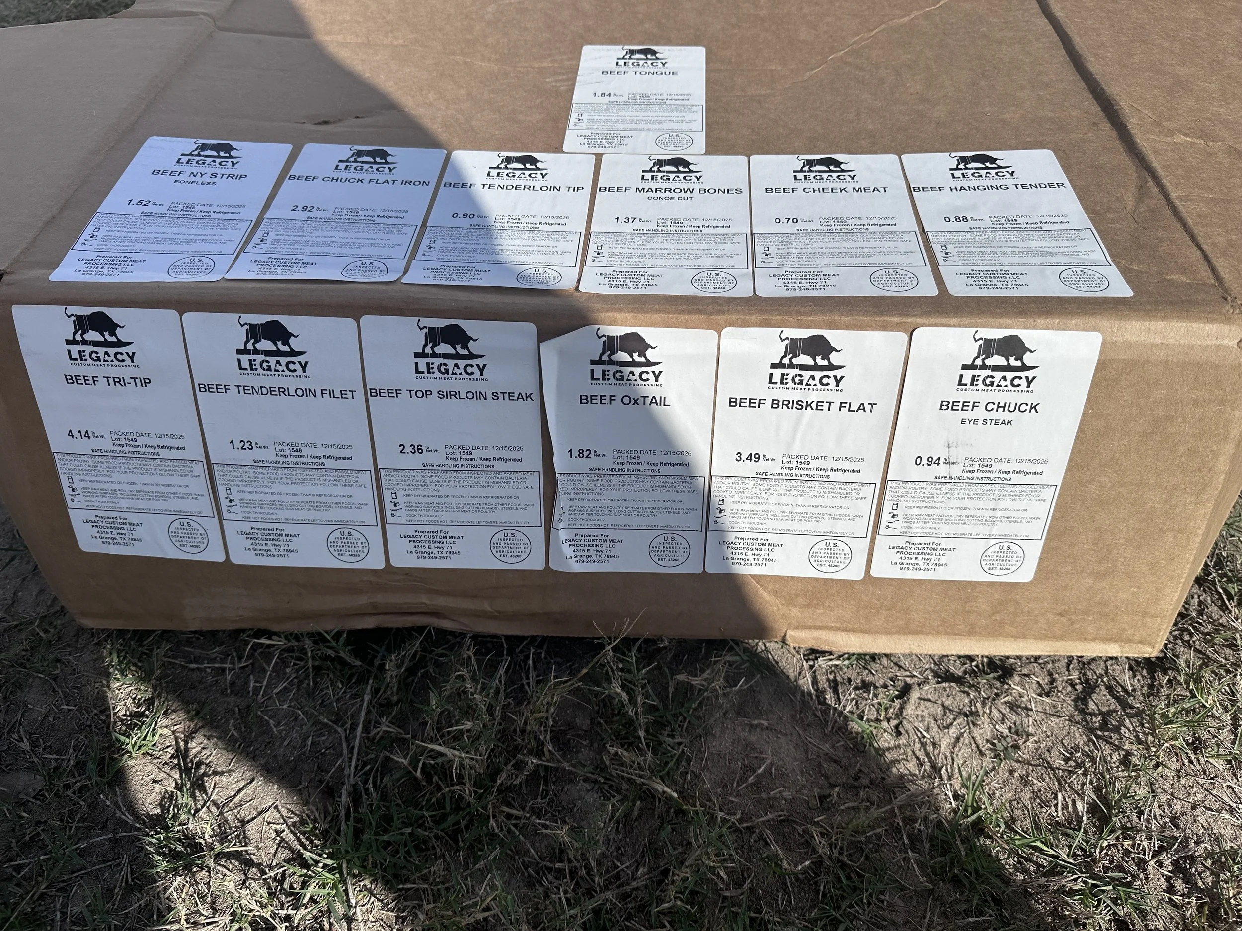 A cardboard box with multiple white labels on top, containing various cuts of beef, placed outdoors on grass and dirt.