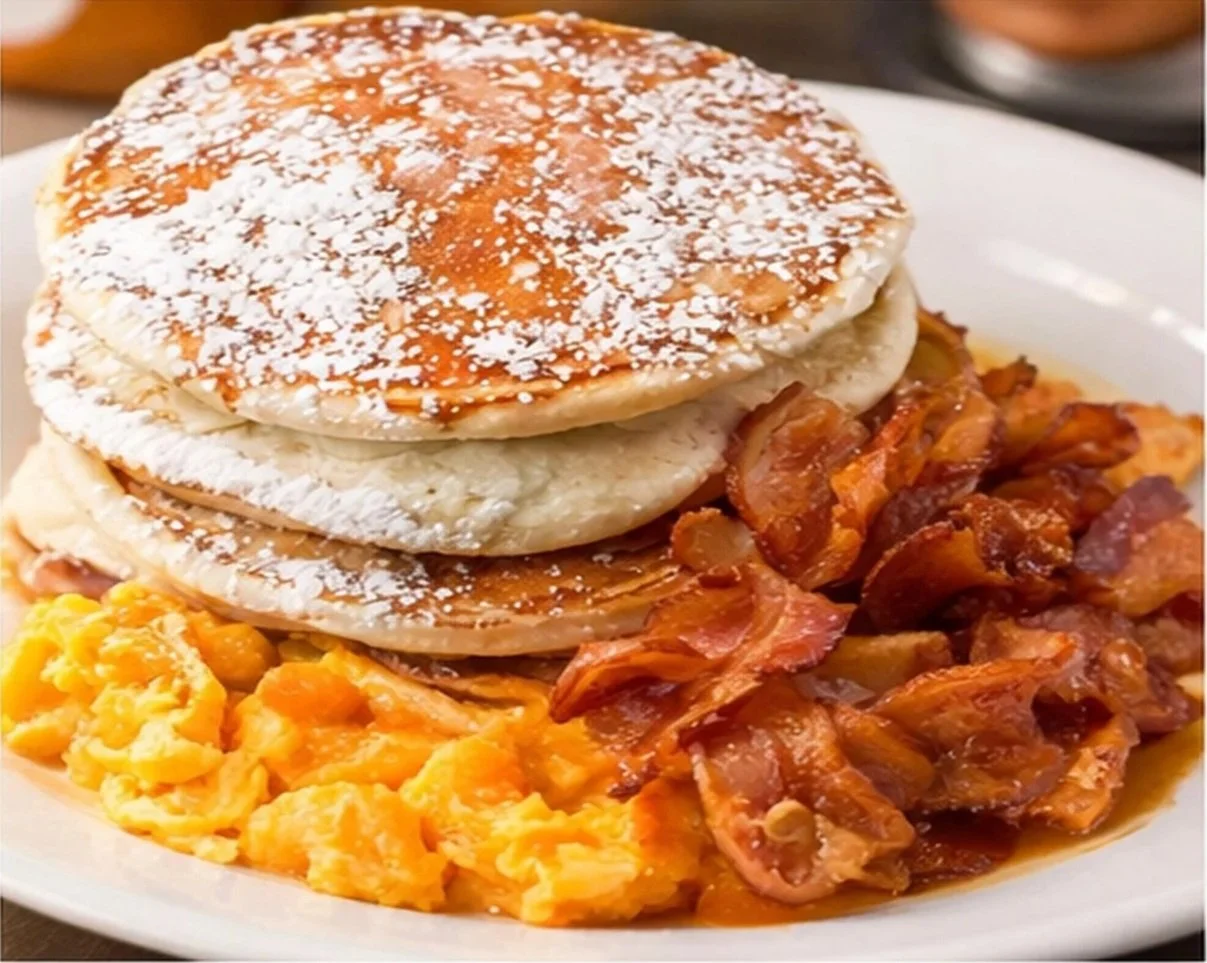 A breakfast plate with toasted English muffins sprinkled with powdered sugar, scrambled eggs, crispy bacon, and sausage gravy with biscuits.