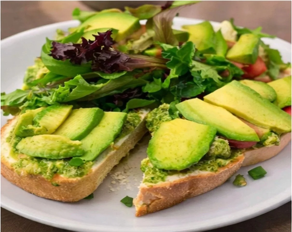 An open-faced avocado toast with mashed avocado topped with sliced avocado on bread, served on a white plate with mixed greens salad.