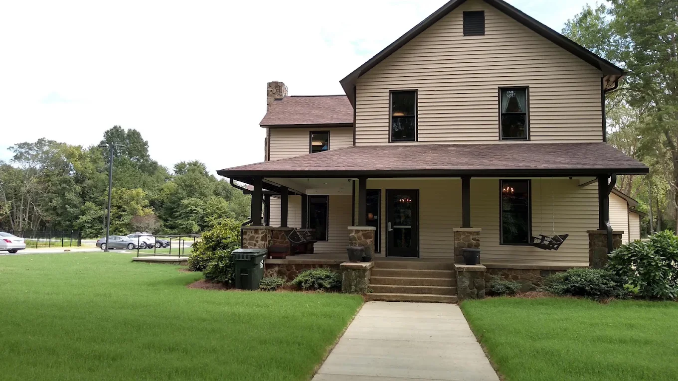 A two-story beige house with black trim, front porch with stone columns and steps, green lawn, trees in the background, and parked cars nearby.