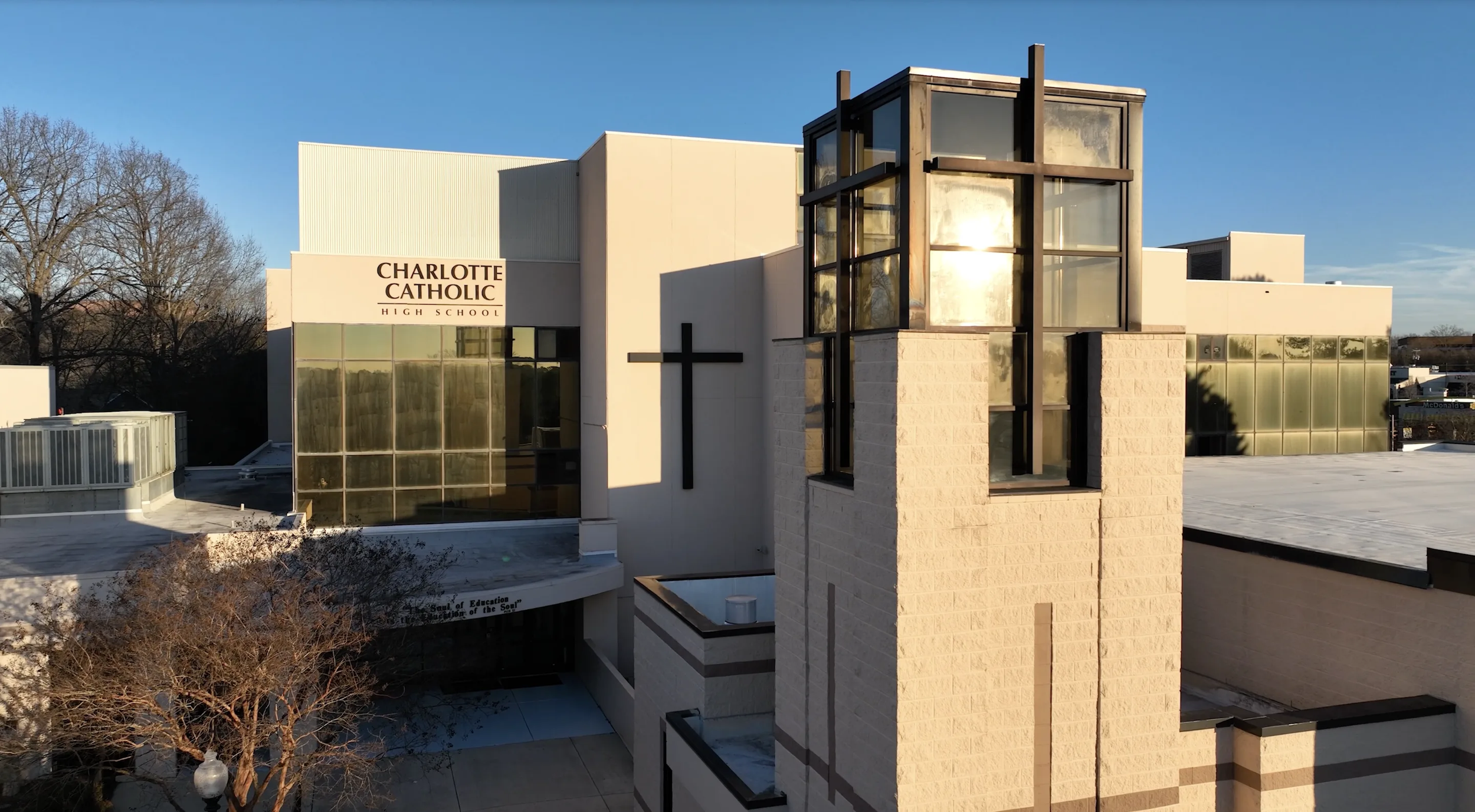 Exterior of Charlotte Catholic High School building with modern architecture, a digital cross, and a glass section reflecting the sunset, under a clear blue sky.