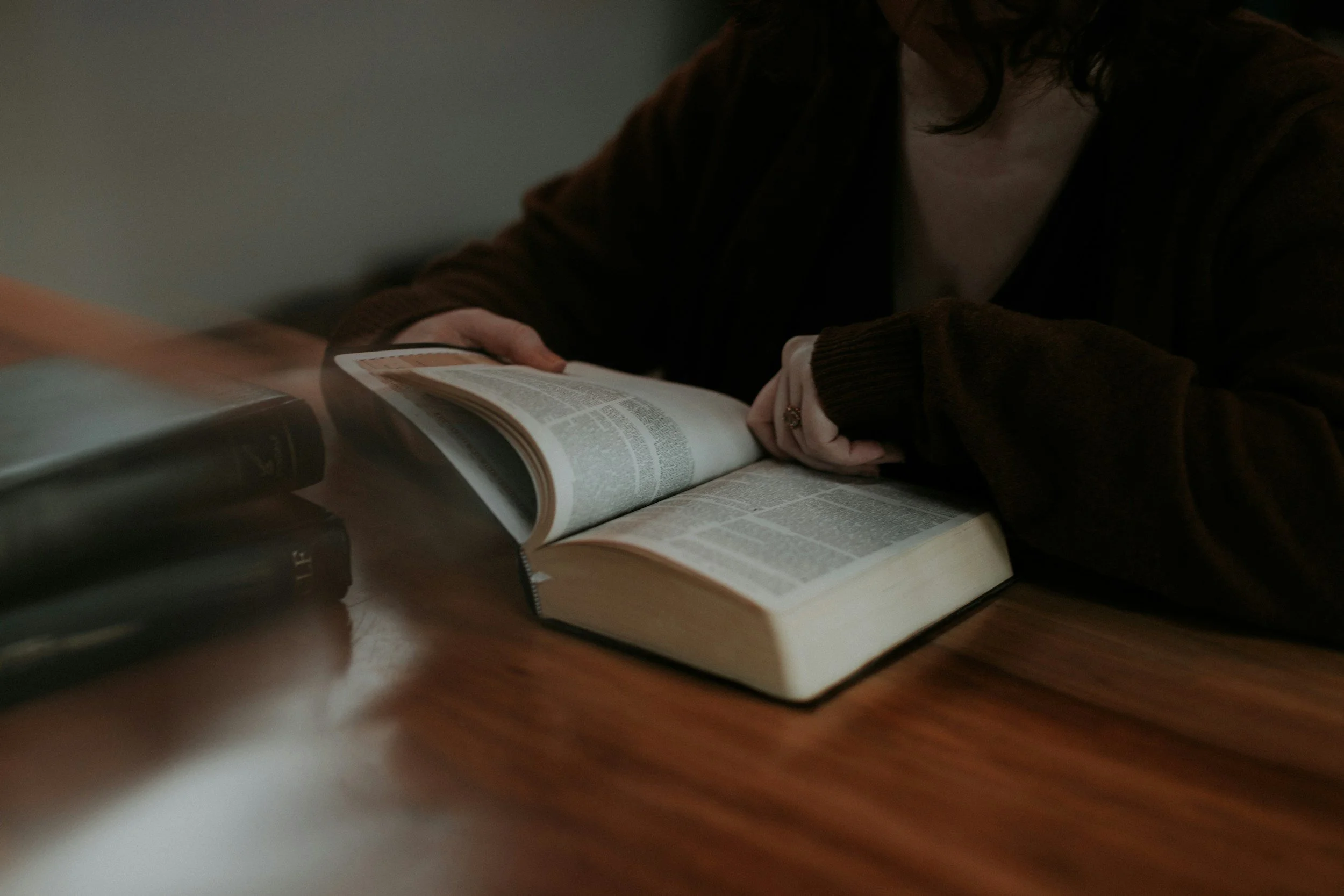 A person with dark hair wearing a dark top, reading a large open book on a wooden table in a dimly lit room.