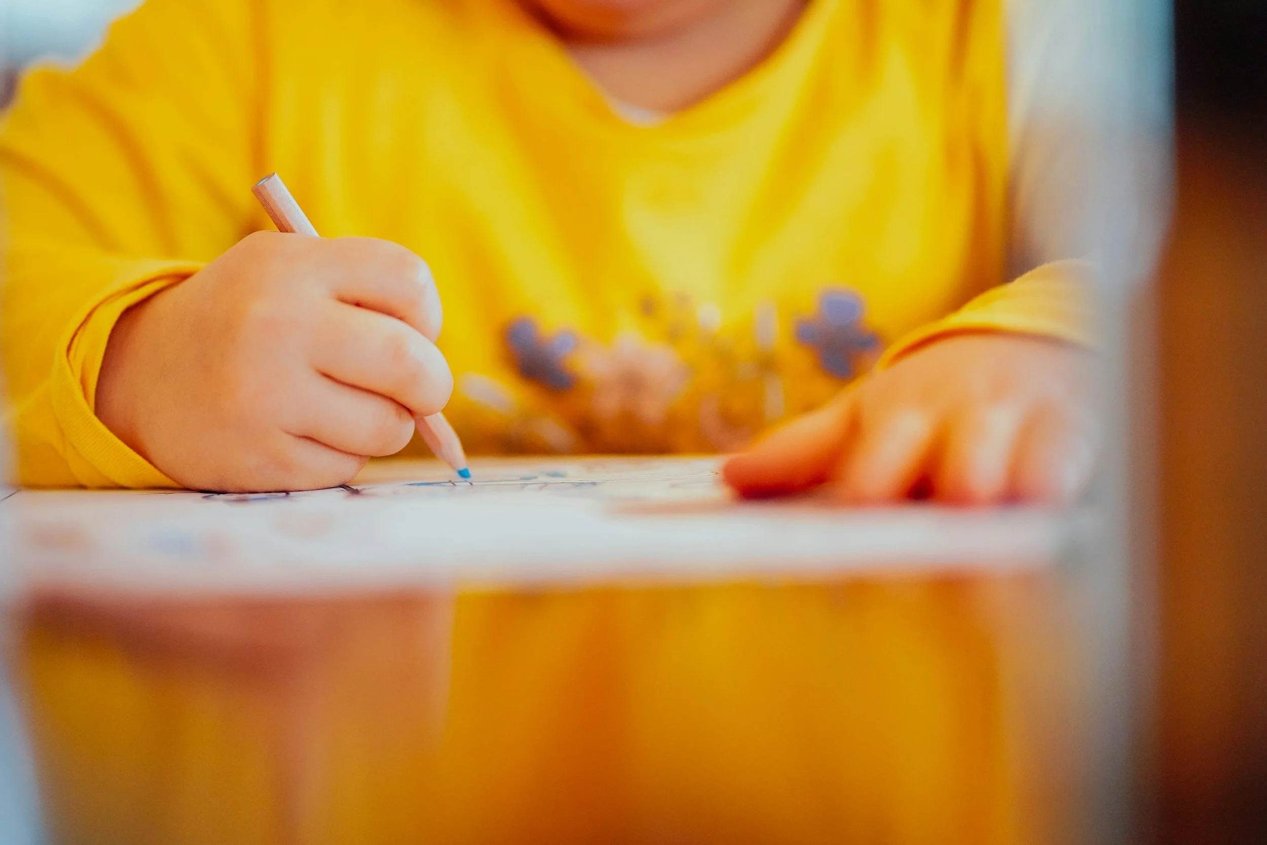 A child's hand holding a crayon and coloring on paper, wearing a yellow shirt.
