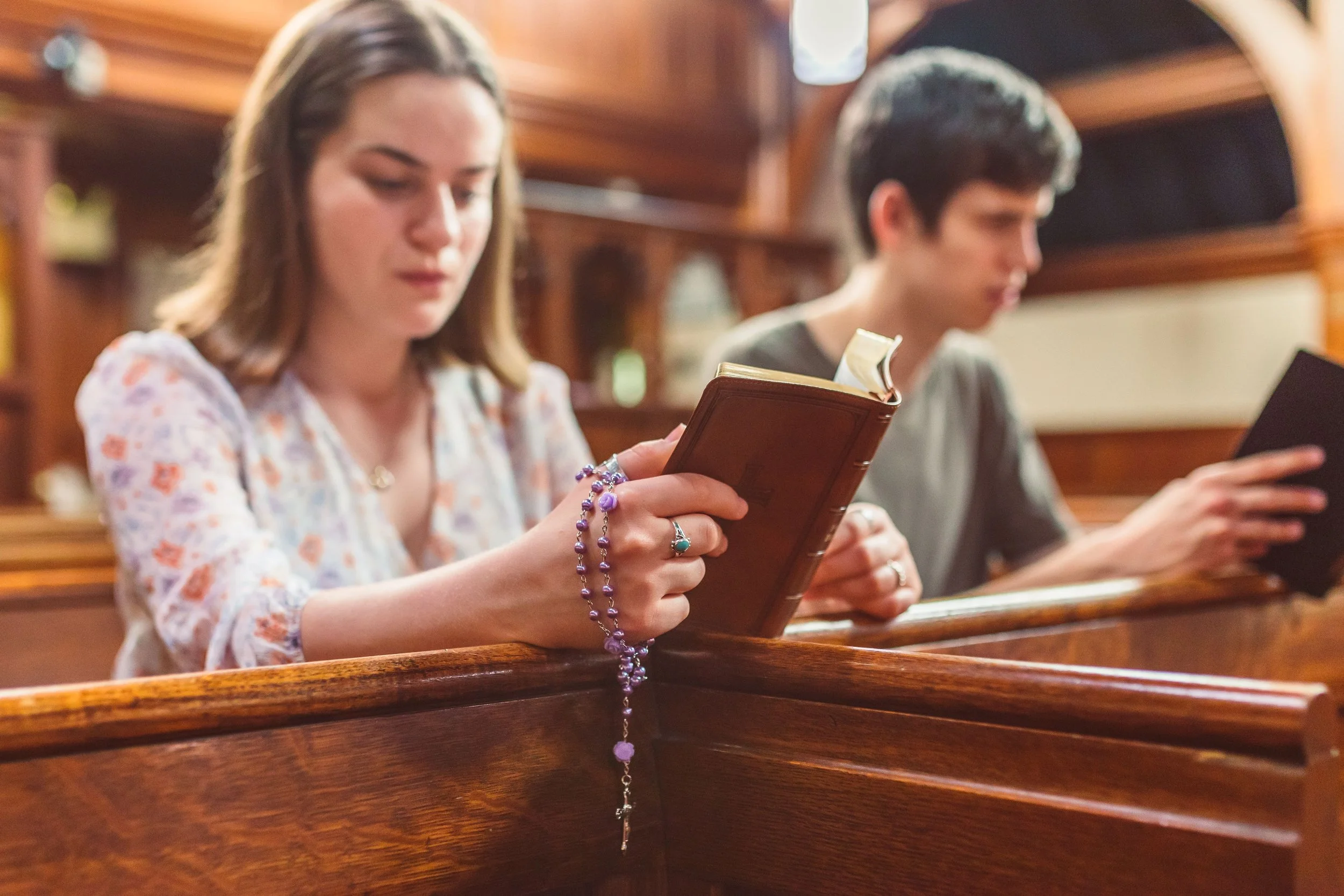 Two people sitting in wooden pews inside a church, reading books or hymnals. The woman in the foreground has light brown hair, wears a floral blouse, and has purple jewelry. The man in the background has dark hair, wears a gray shirt, and is focused on his book.