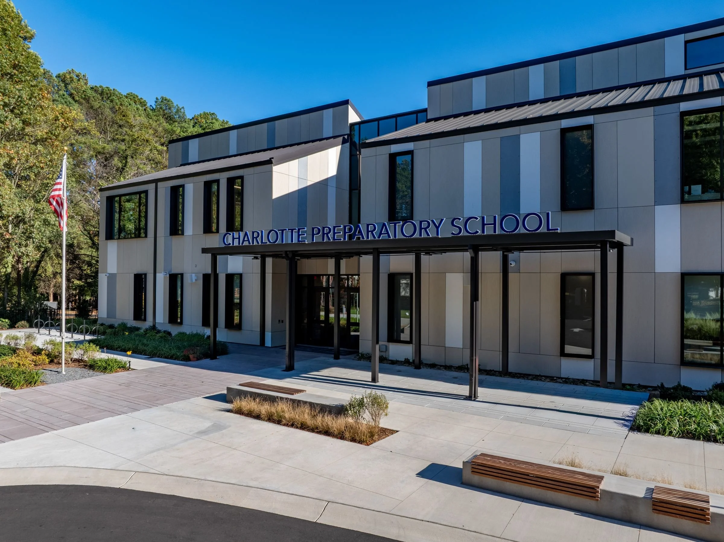 Modern school building with a sign that reads Charlotte Preparatory School, surrounded by trees and a paved outdoor area with benches and landscaping.