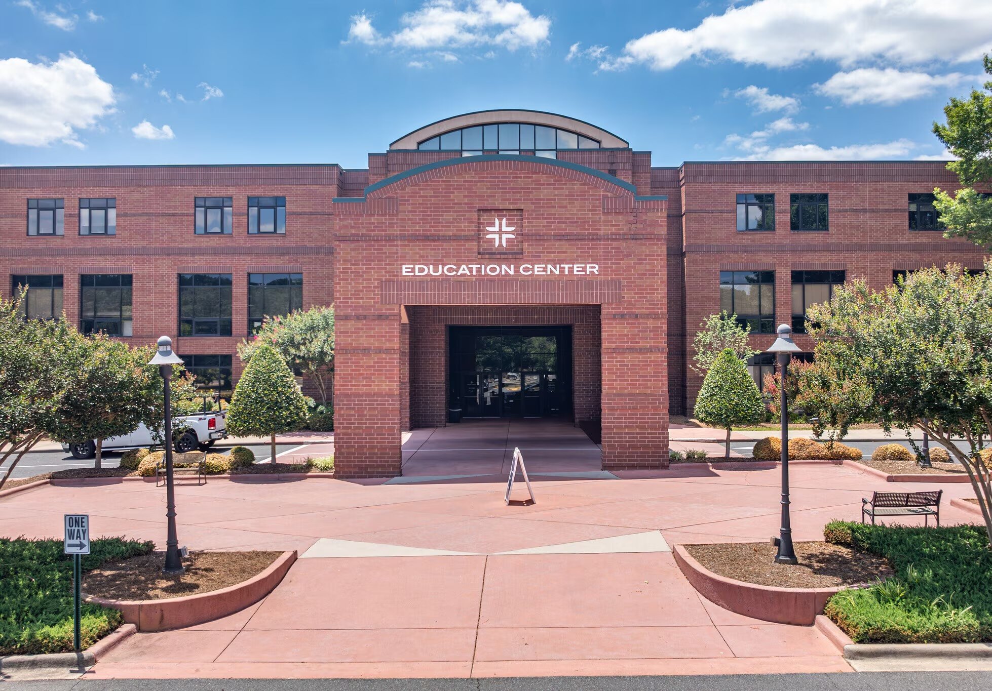 Front view of a brick education center building with a large glass window arch on top, surrounded by trees, parked cars, and a pink sidewalk, under a blue sky with scattered clouds.
