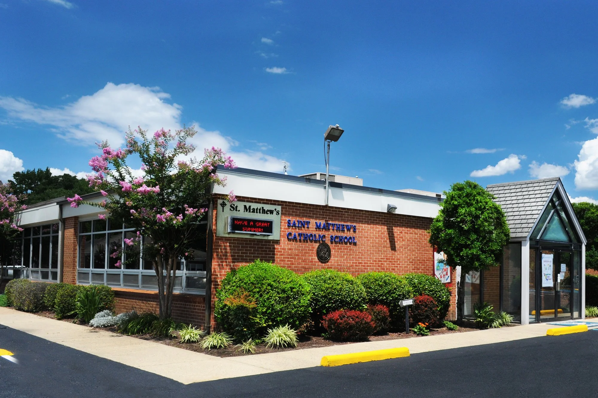 The exterior of Saint Matthew's Catholic School building with a sign reading 'Have a Great Summer!', a flowered tree, green bushes, a glass door entrance, and a blue sky with clouds.