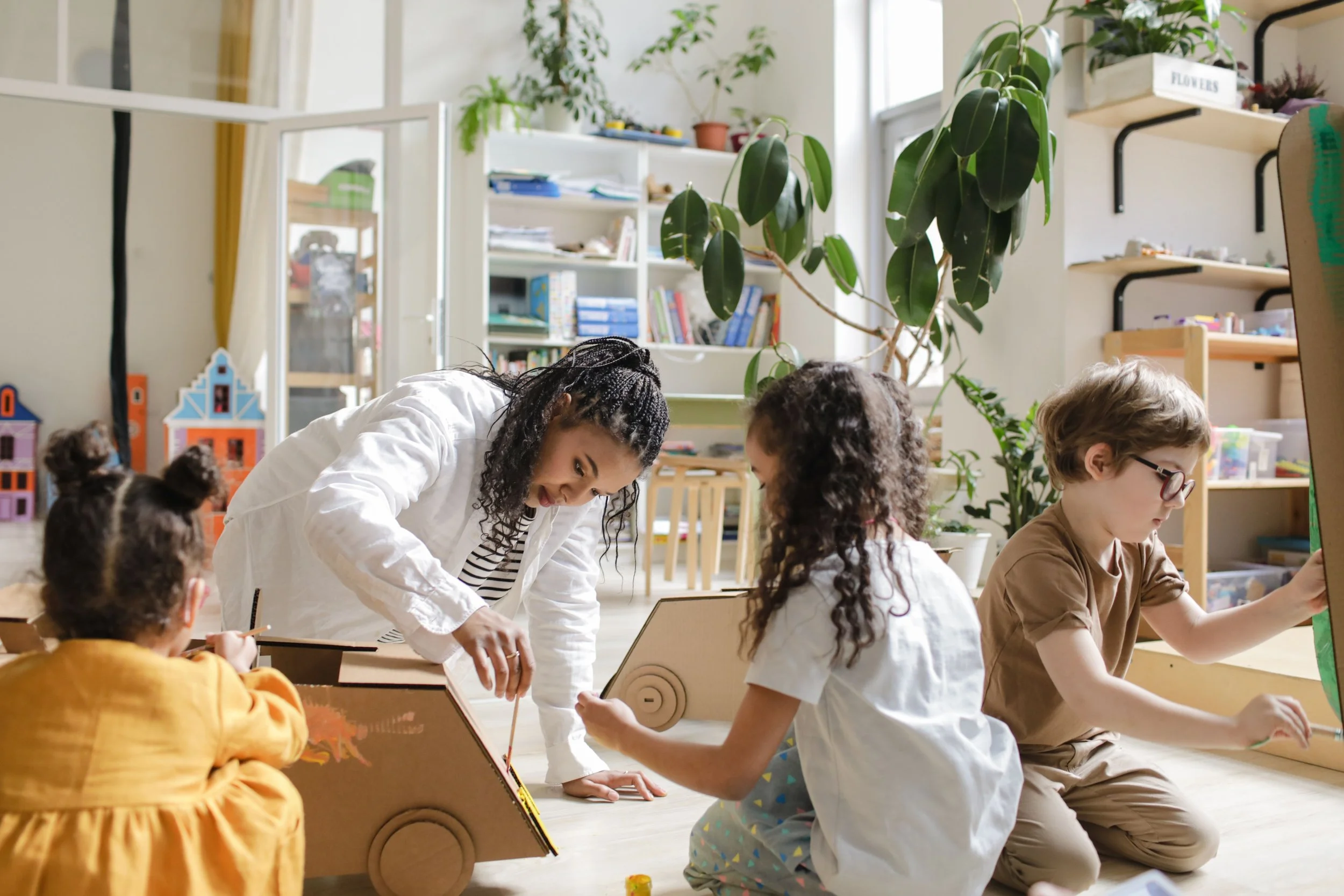 A teacher interacting with children in a classroom filled with books and plants.