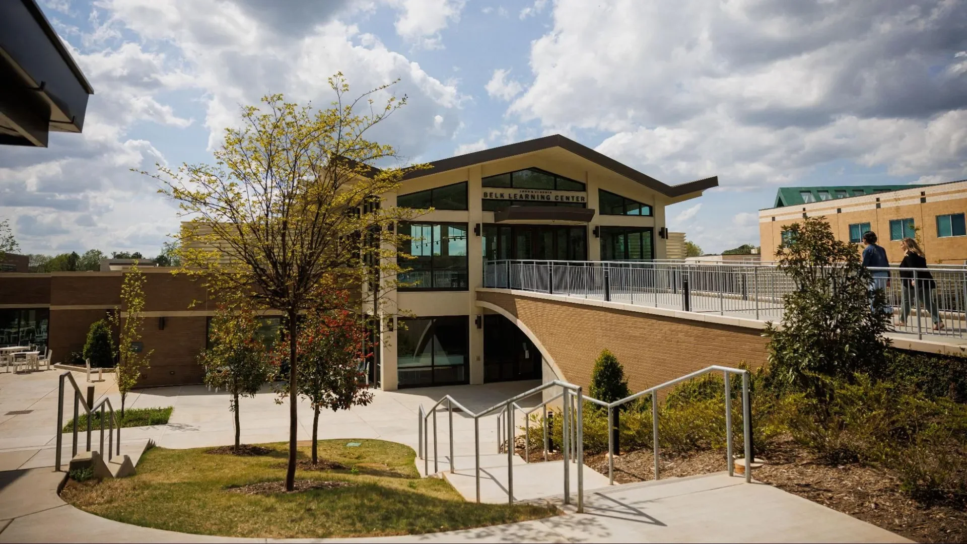 Exterior view of the Belk Learning Center building on a sunny day with a partly cloudy sky, trees, and people walking on the walkway.