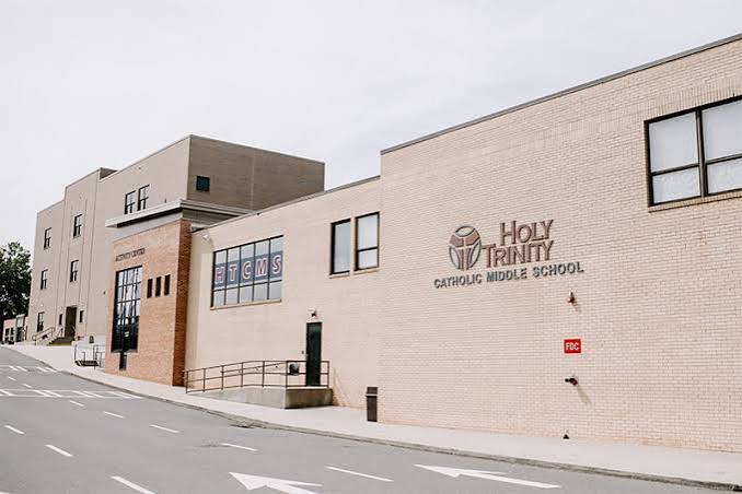 Exterior view of Holy Trinity Catholic Middle School building on a sloped street with road markings, brick and beige walls, and windows.