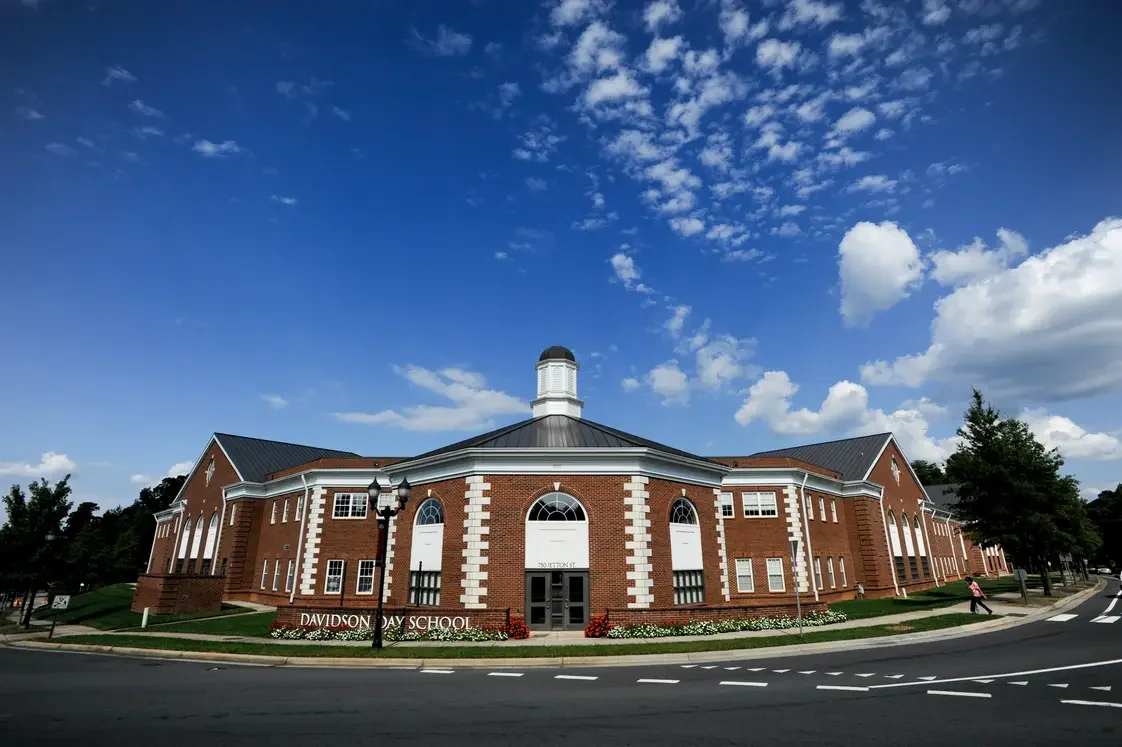 A large brick school building with white accents and arched windows, labeled Davidson Day School, under a blue sky with scattered clouds.