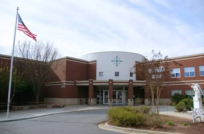 Front view of a church with a cross above the entrance, a flagpole with the American flag, and some trees and shrubs in front.