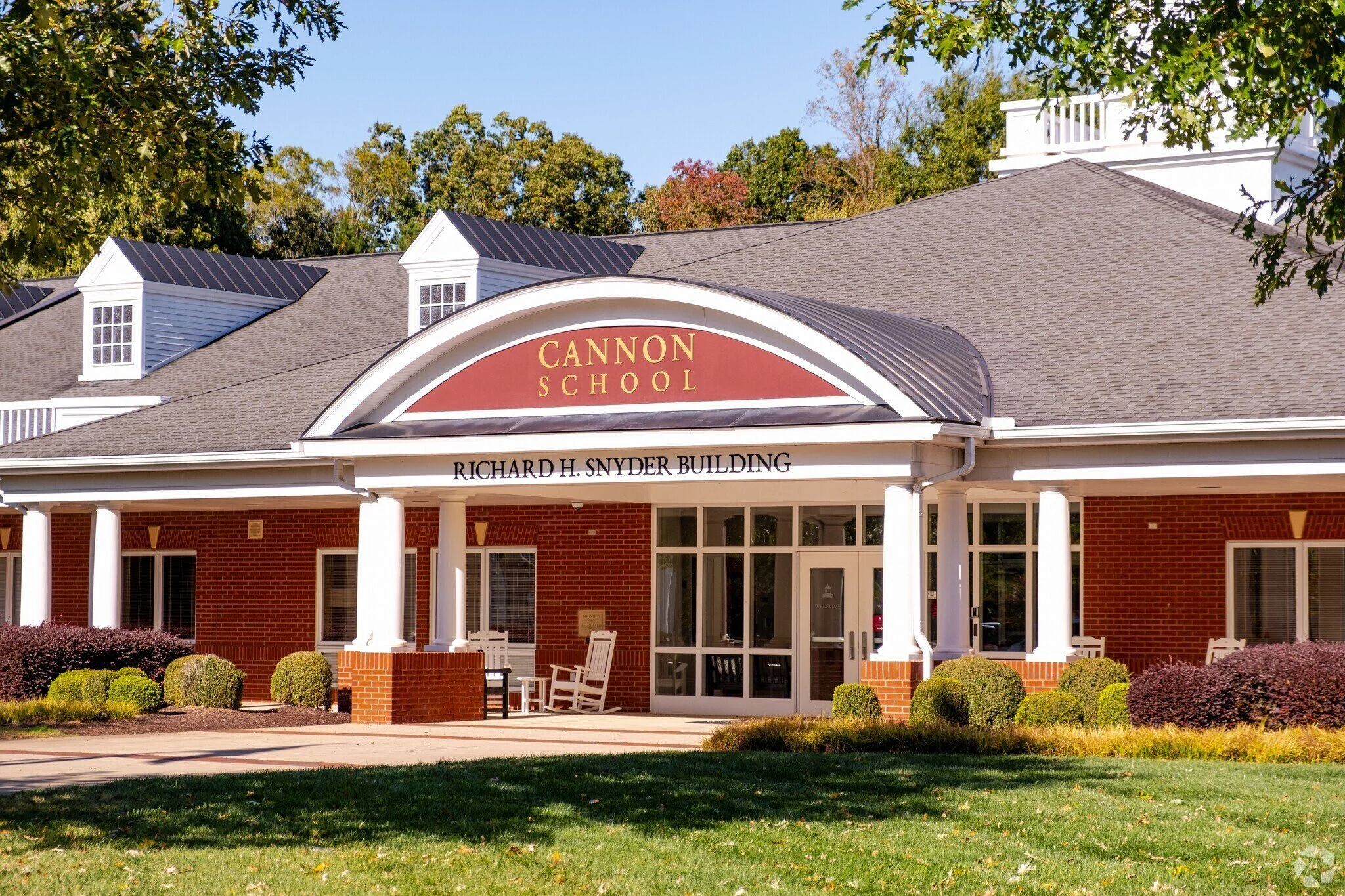 Front exterior of Cannon School, Richard H. Snyder Building, with brick walls, white columns, and a gabled roof, surrounded by trees and shrubs.