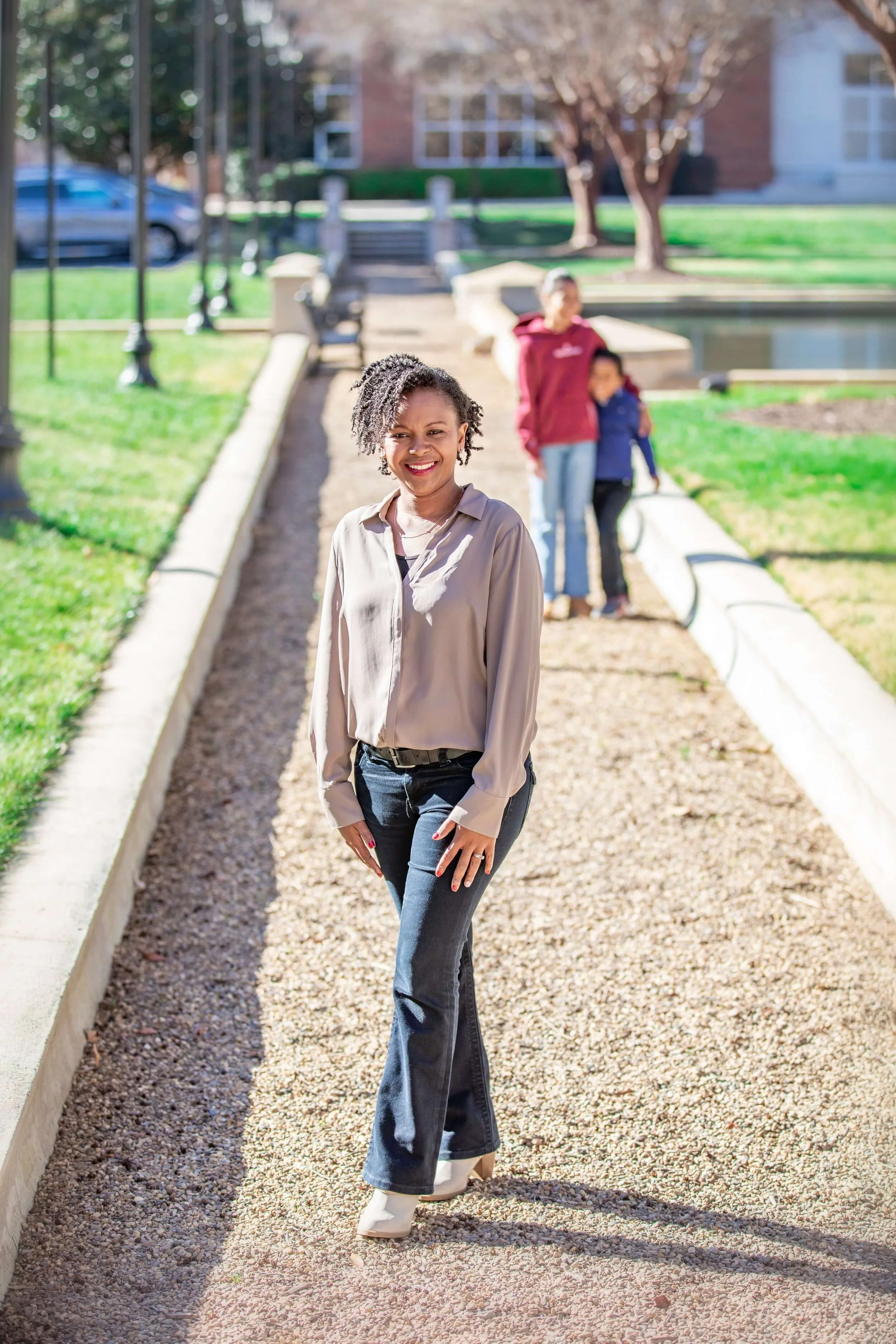 Woman with curly hair smiling, standing on a gravel walkway outdoors, with two people blurred in the background near a small pond and trees.