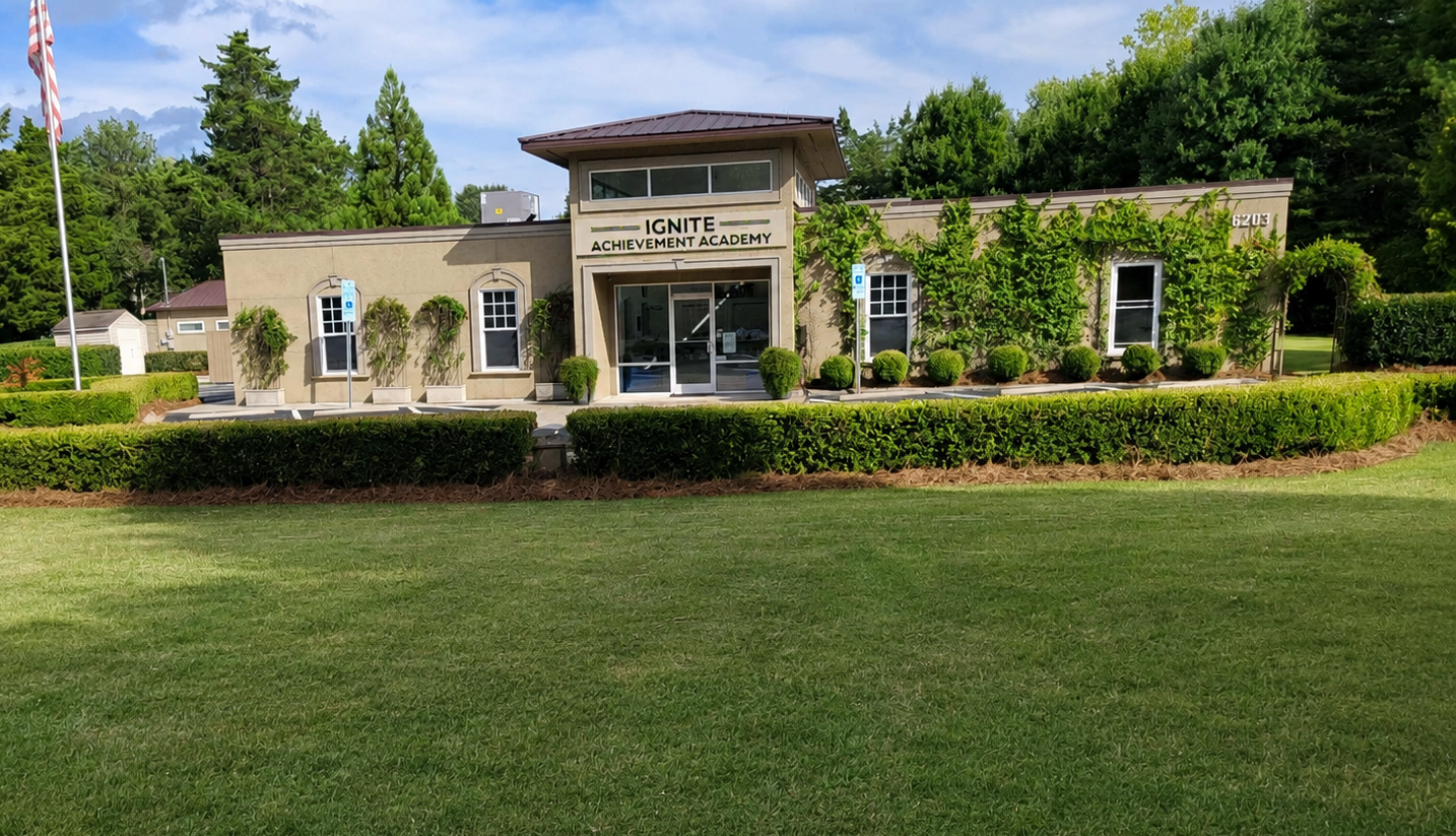 A single-story building labeled Ignite Achievement Academy with a tan facade, surrounded by green bushes and trees, and a parking lot visible to the sides.