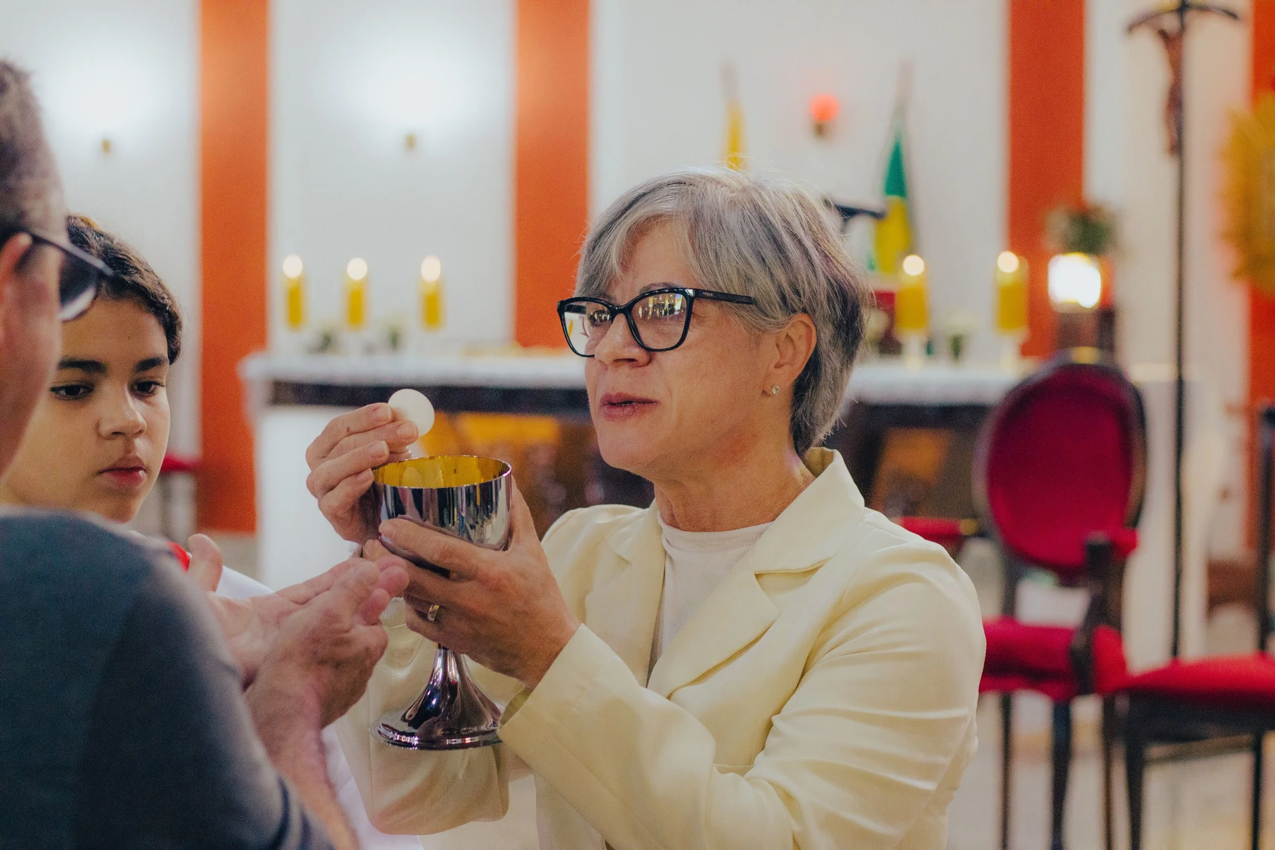 An elderly woman with gray hair and glasses holding a chalice, participating in a religious ceremony with a young man and other individuals nearby inside a decorated church.
