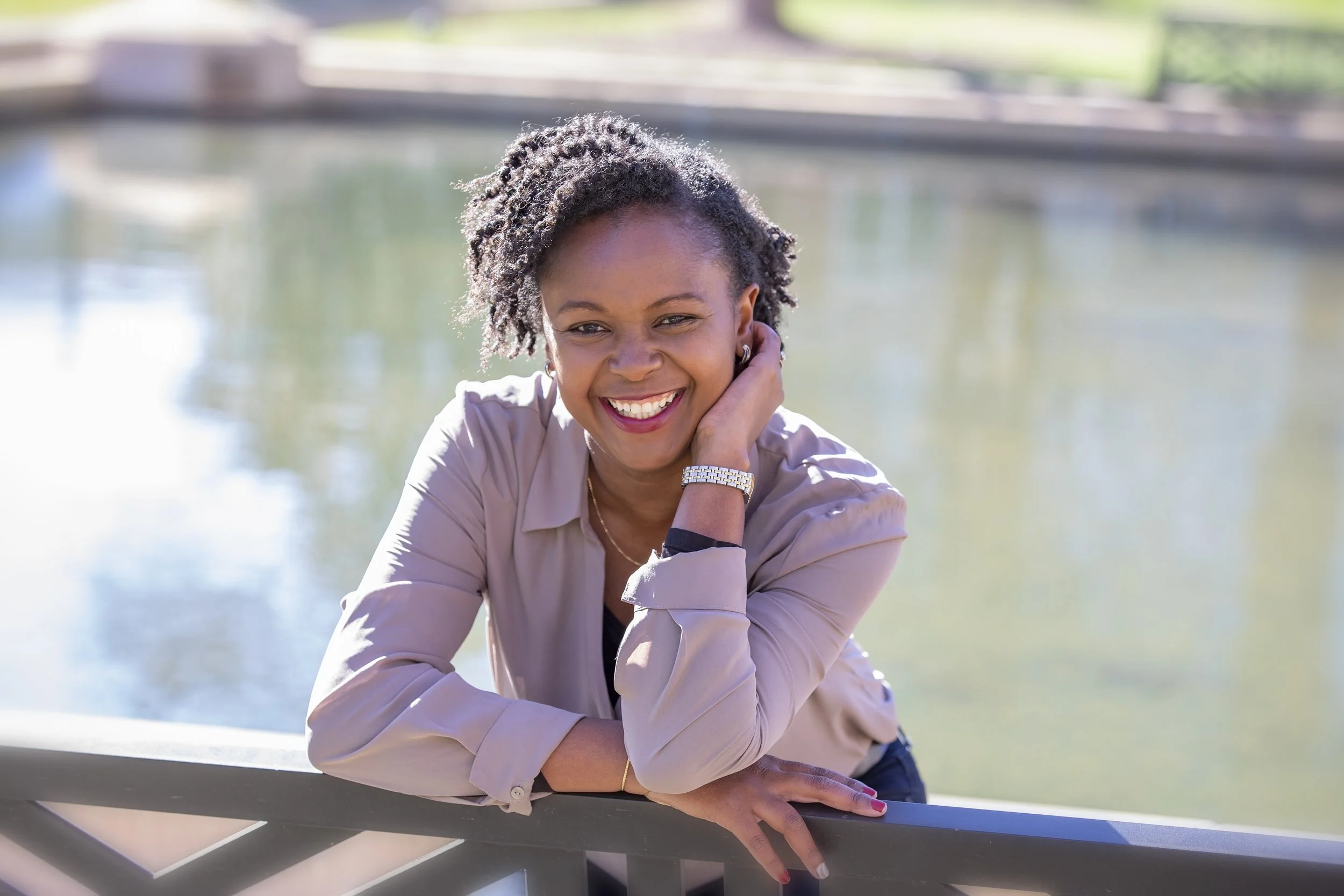 A woman with curly hair smiling, leaning on a railing outdoors near a body of water, wearing a beige jacket and gold watch.