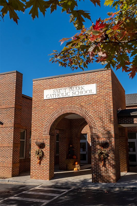 The entrance to Saint Mark Catholic School with a brick archway and a sign above reading 'Saint Mark Catholic School.' There are fall decorations and a tree with colorful leaves overhead.
