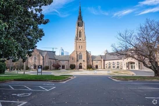 A church with a tall steeple surrounded by trees and buildings, under a blue sky.