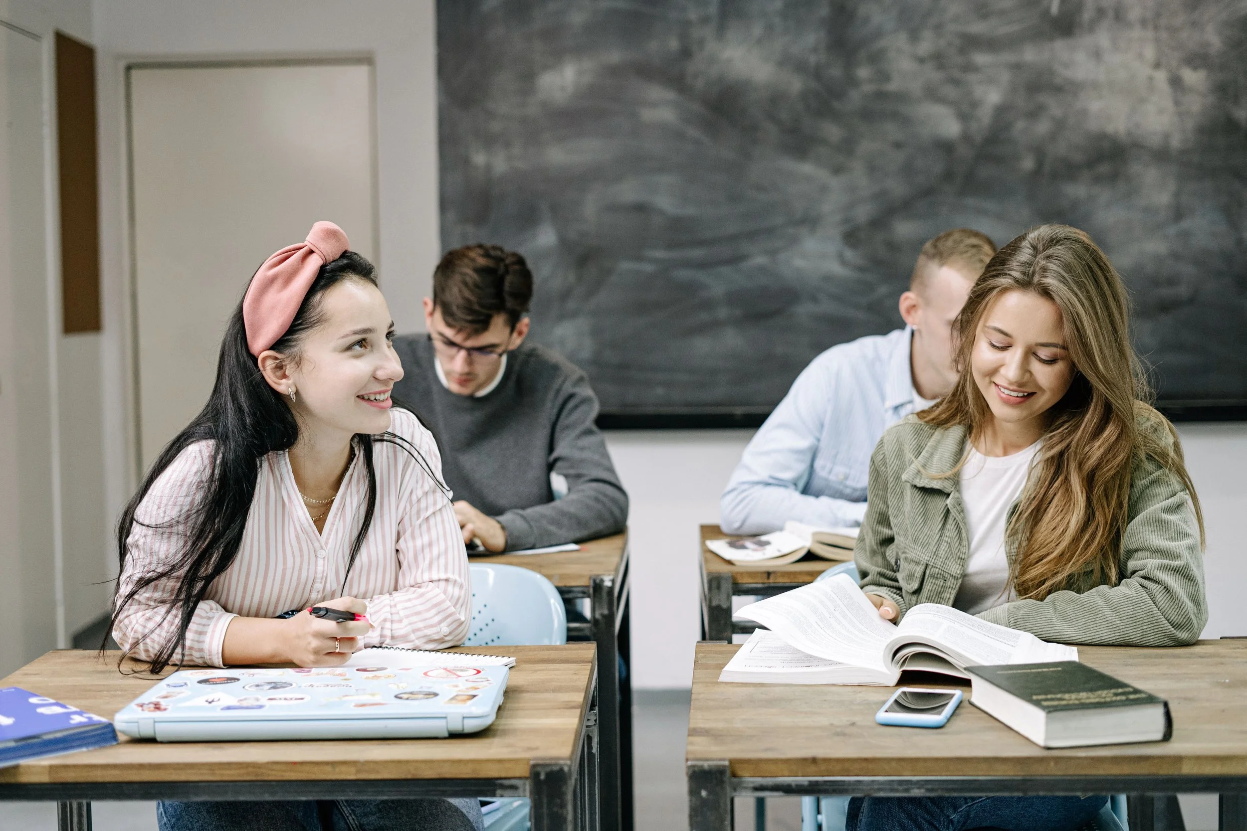 Four students sitting at desks with open books and notebooks in a classroom, two female students in the foreground smiling and engaged, one with long dark hair and a pink headband, the other with wavy blonde hair. Two male students in the background reading.