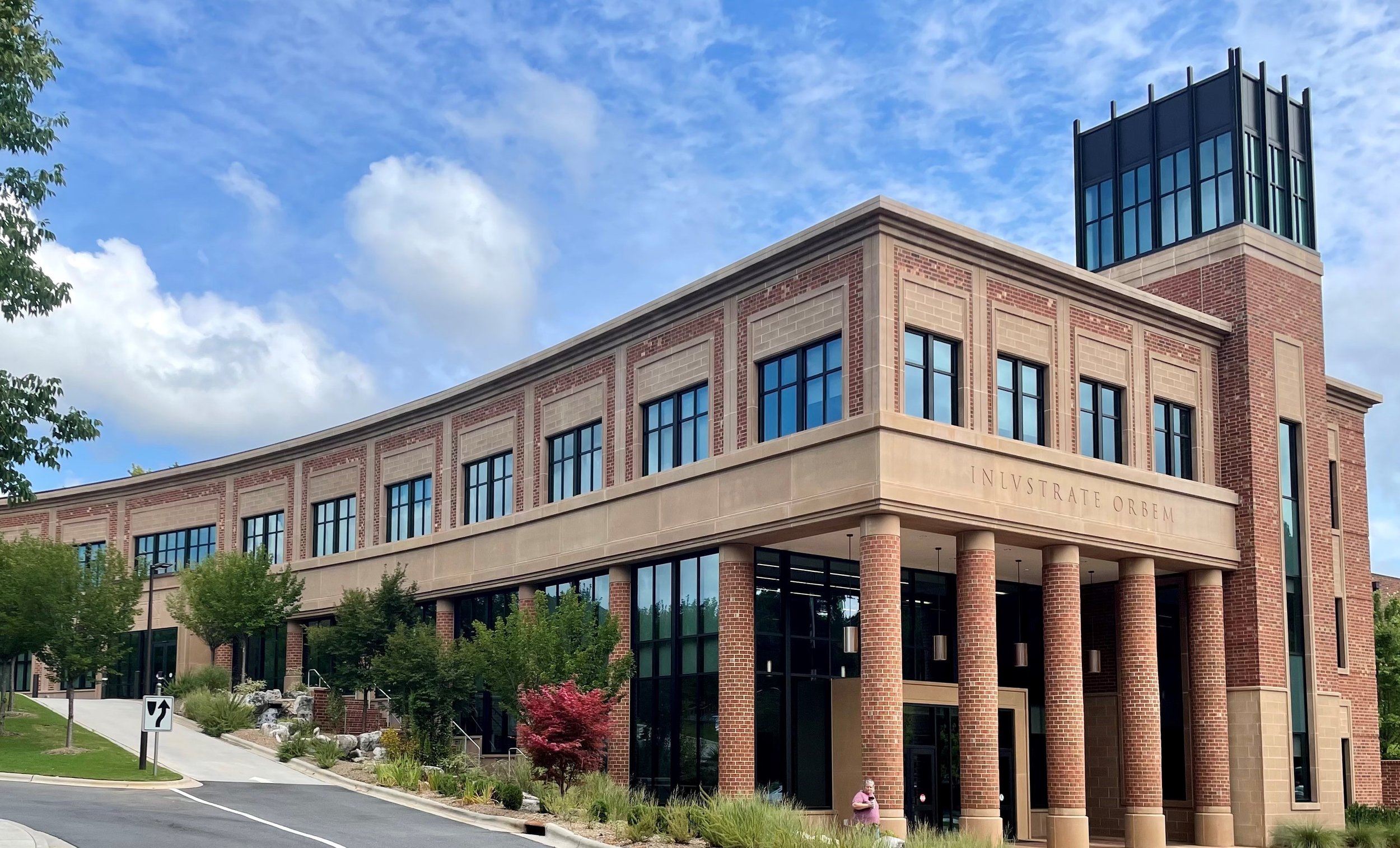 A modern brick and glass building with columns, trees, and a person walking near the entrance against a partly cloudy sky.