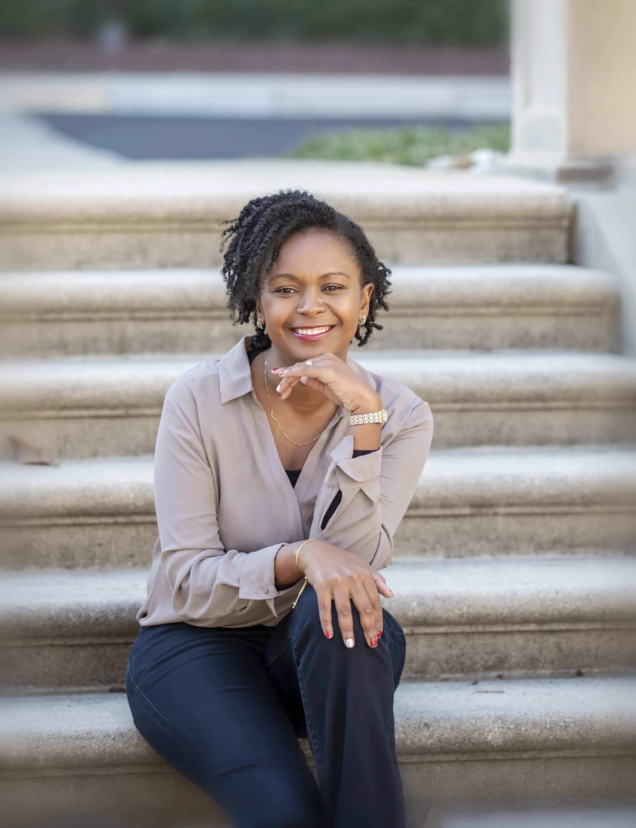 A woman sitting on outdoor steps, smiling and resting her chin on her hand, wearing a beige blouse, dark blue jeans, earrings, necklace, and a watch, with a blurred background of grass and concrete.