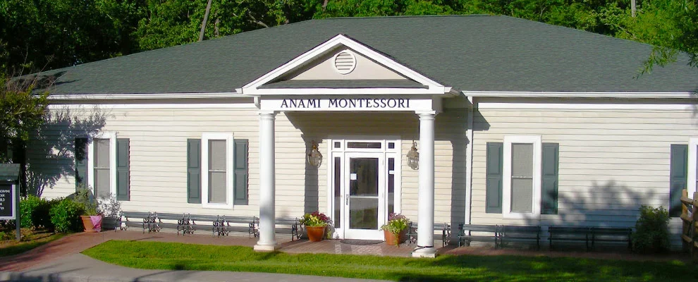 Exterior of Anami Montessori school building with white siding, gray trim, four windows with shutters, and a front entrance supported by two columns.