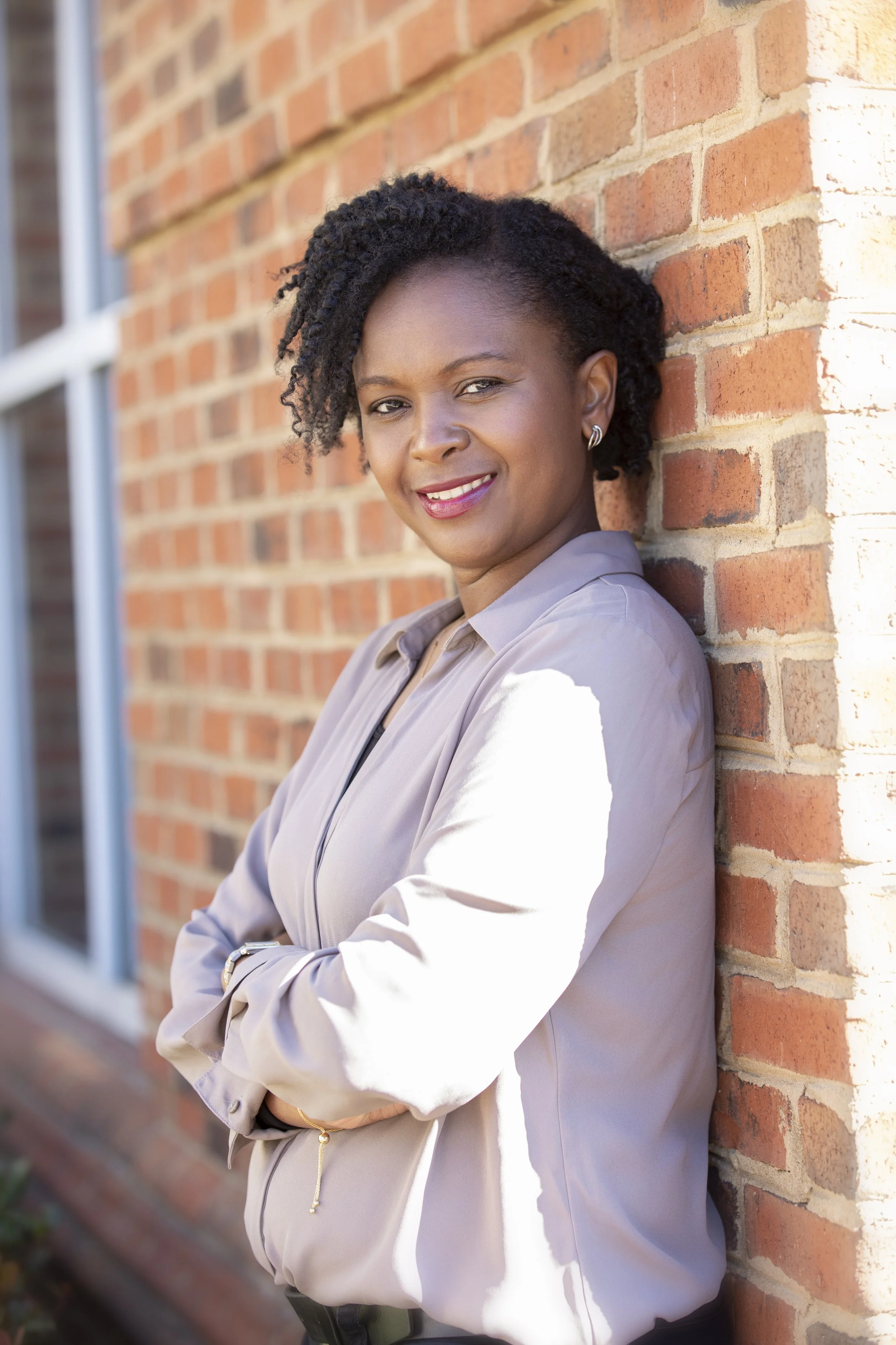 A woman with dark, curly hair smiling while leaning against a brick wall outdoors.
