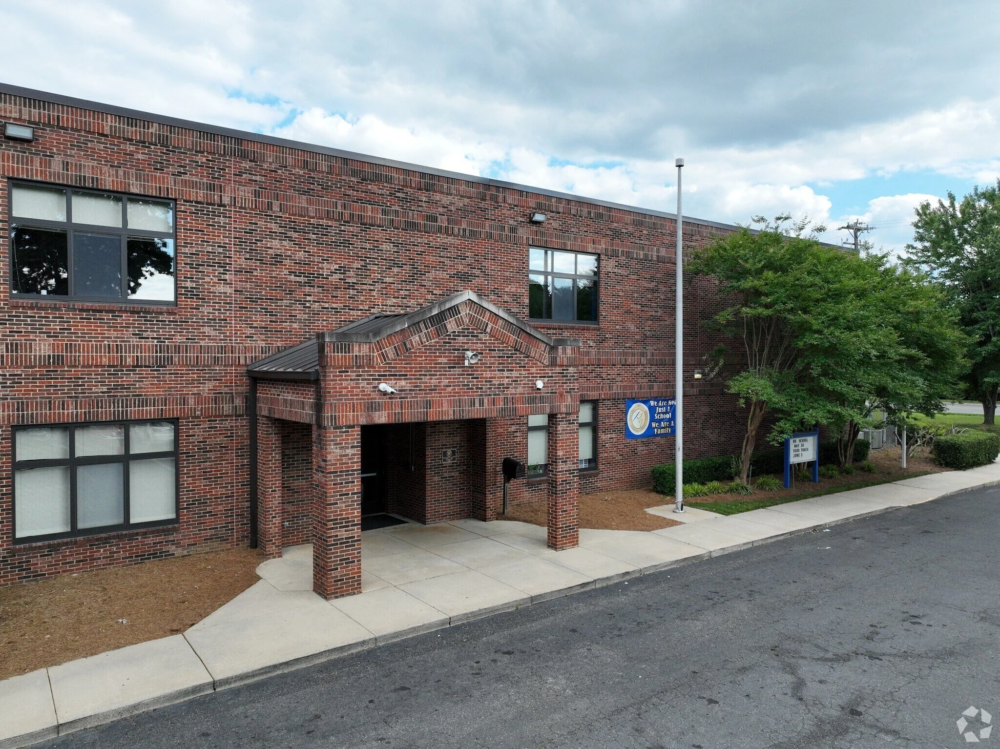 Brick school building with multiple windows, trees, and informational signs in the foreground under a partly cloudy sky.