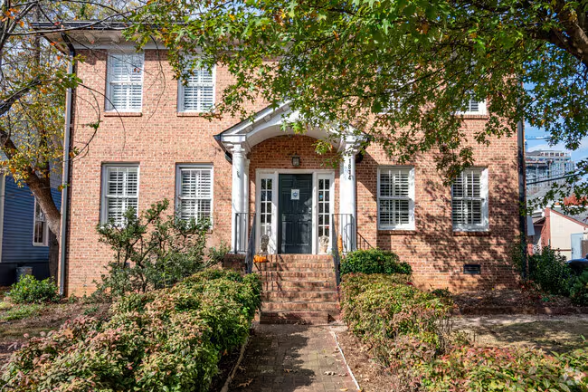 A brick two-story building with a white arch over the front door, a small set of stairs, and several windows with shutters. There are trees and bushes in front of the building.